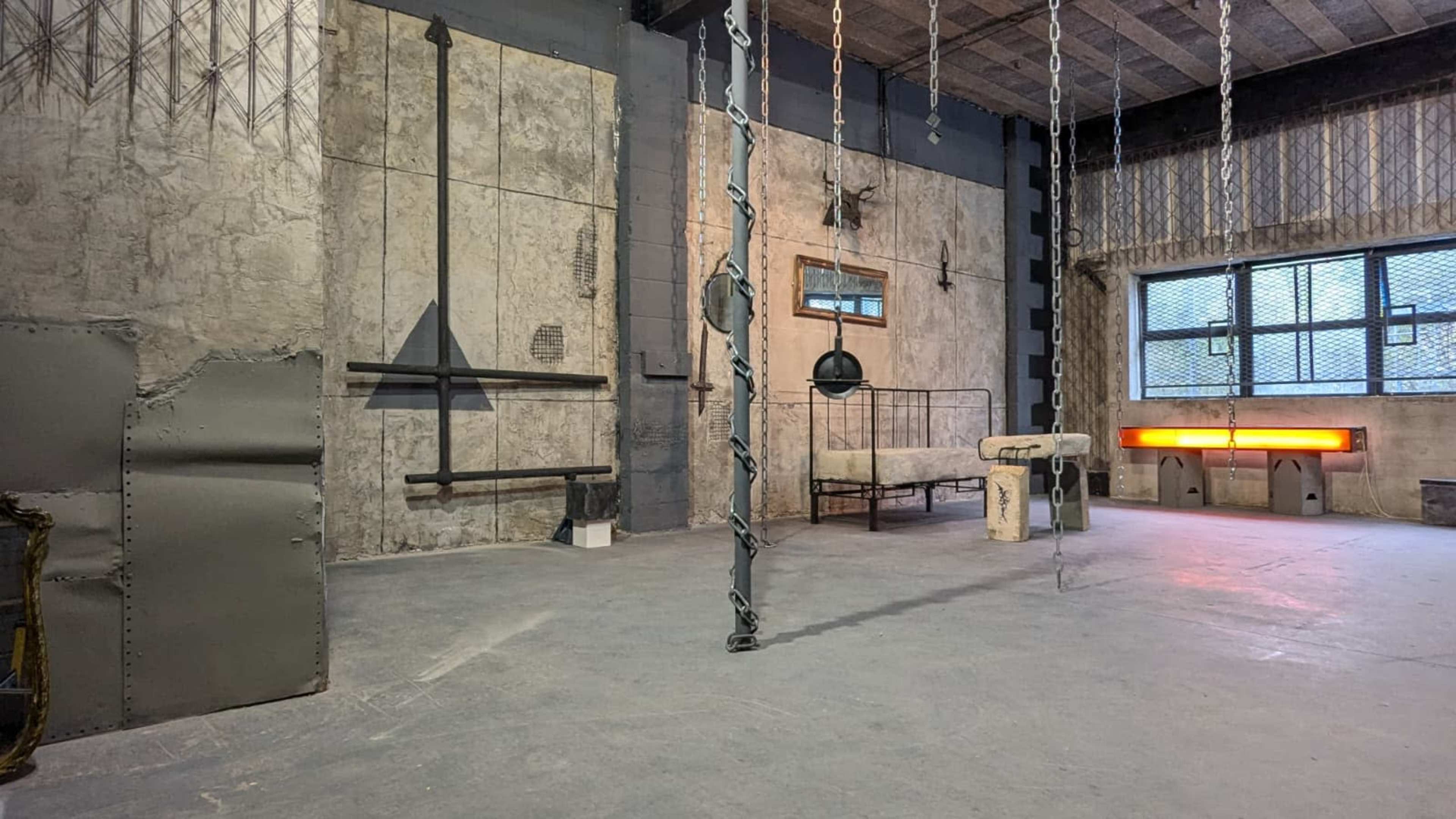 The image shows an industrial-style room with concrete walls, a gray floor, and various metal structures including chains, a suspended object, and an illuminated panel.