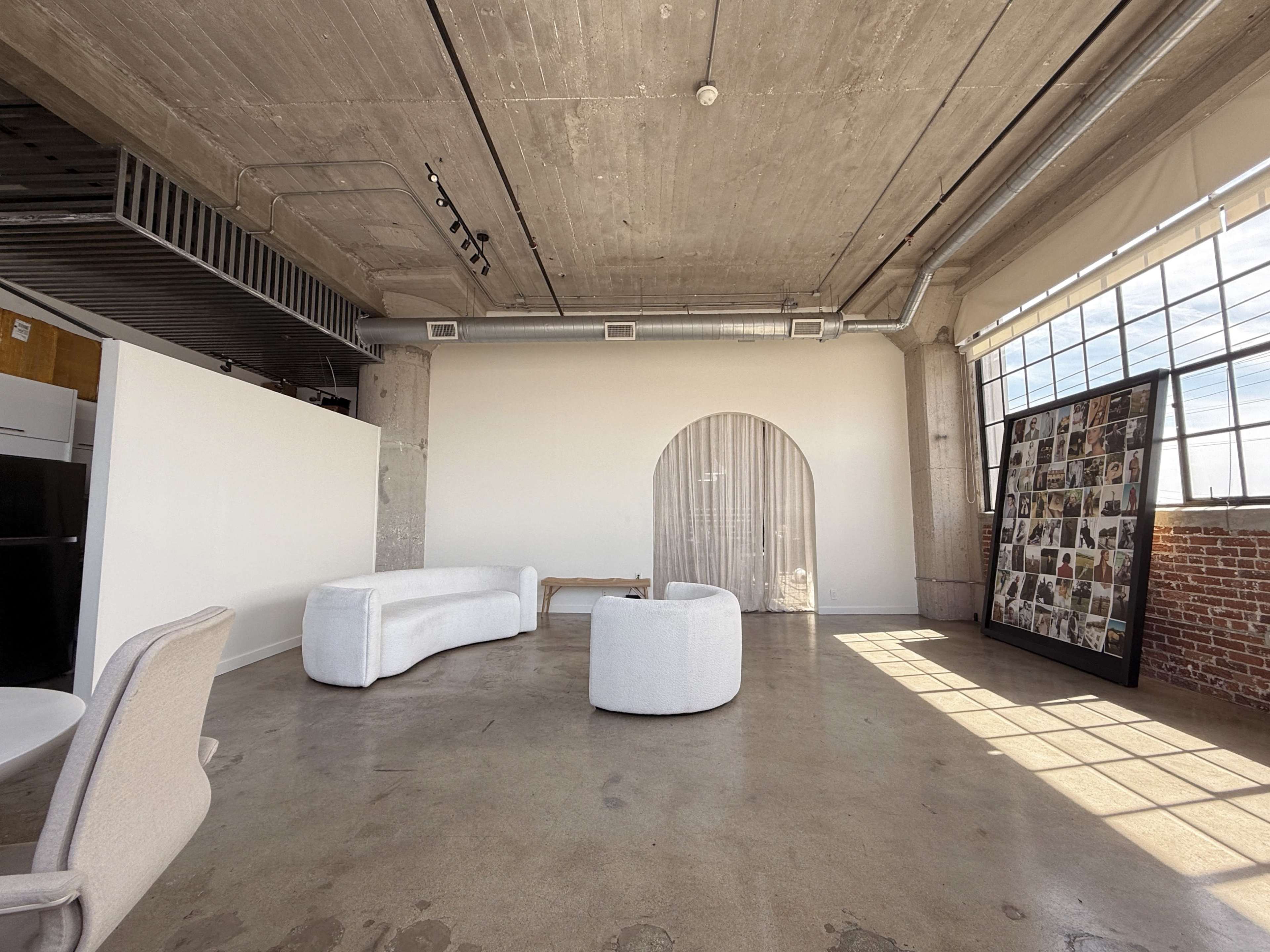 The image shows a spacious modern interior with concrete walls, a large window allowing natural light, and minimal furniture including a curved sofa and an armchair.