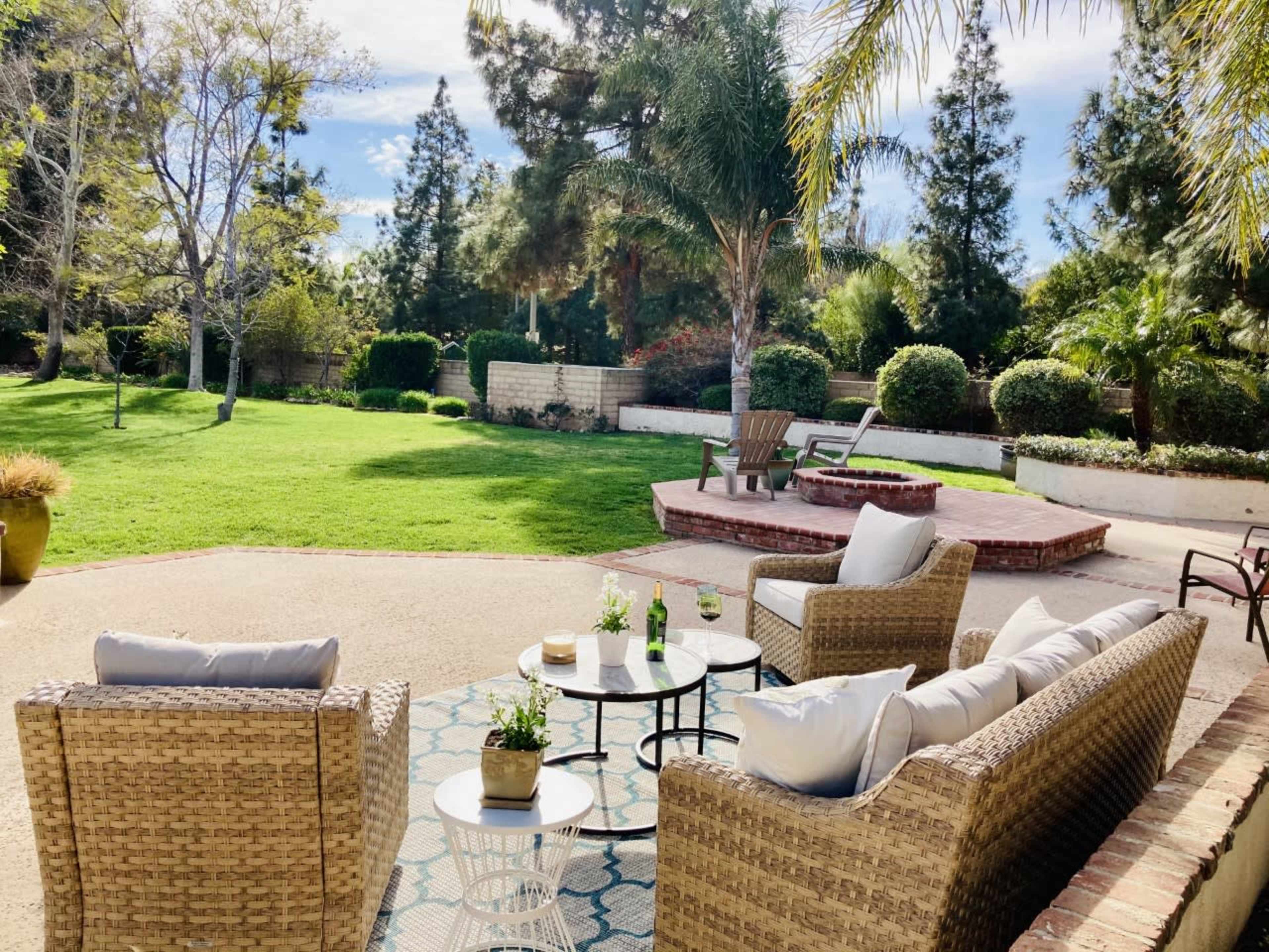 A patio area with wicker furniture and a small table overlooks a green lawn surrounded by trees and shrubs.