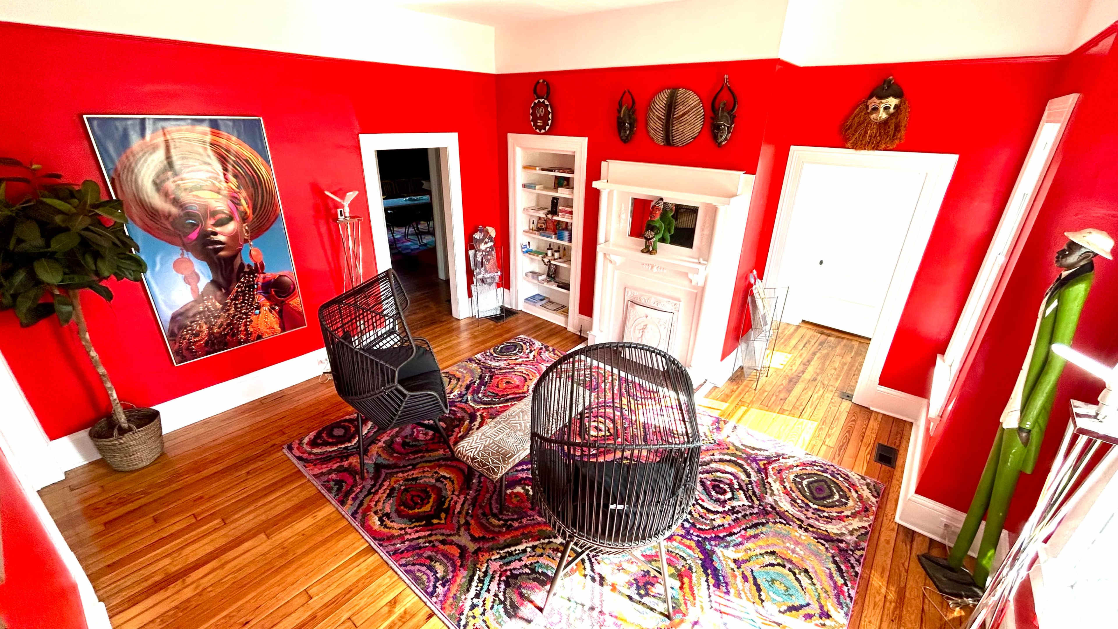 The image shows a brightly colored red room featuring two black chairs, a colorful rug, and various decorative items on the walls and shelves.