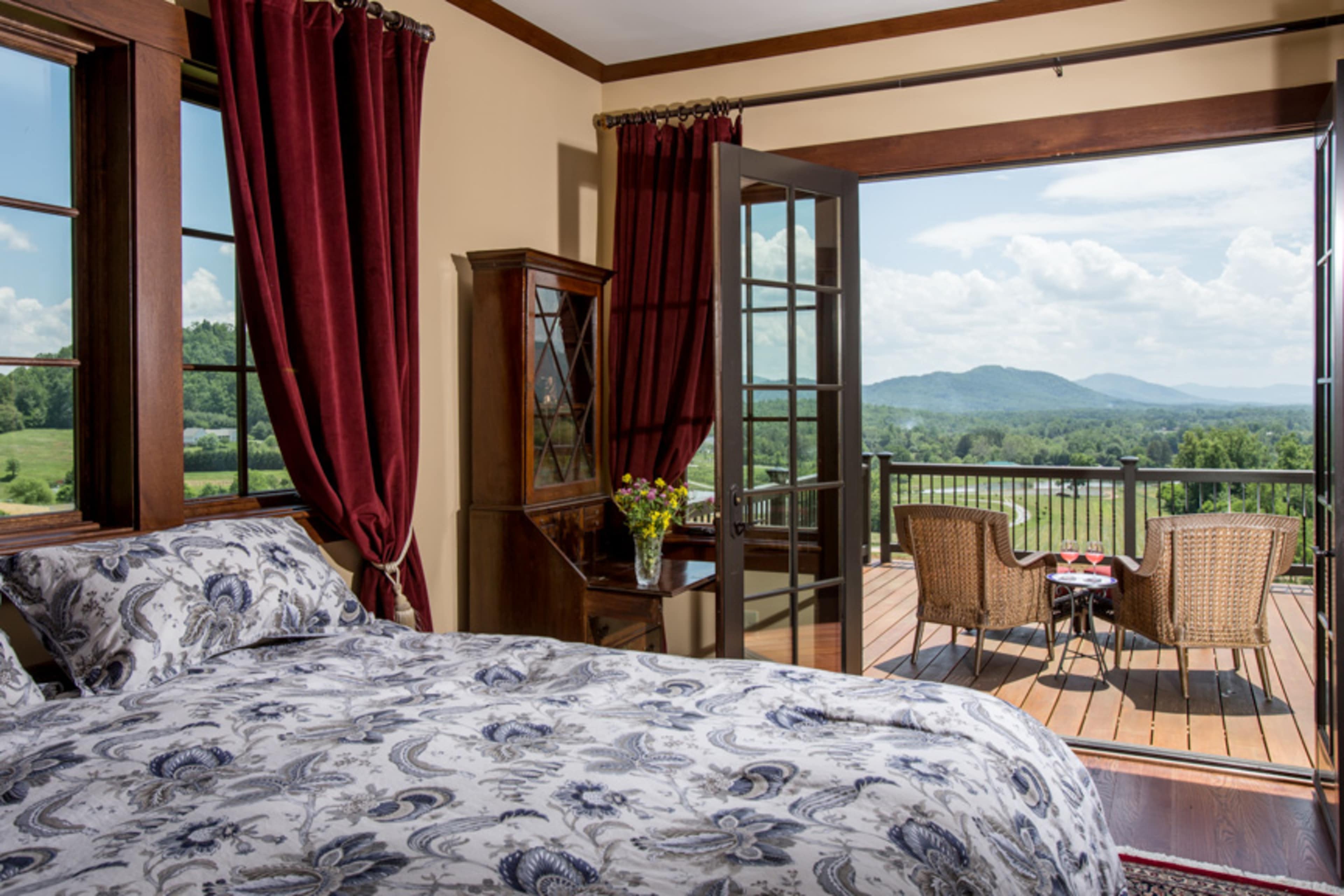 A spacious bedroom features a bed with patterned bedding, large windows with red curtains, and a balcony that opens to a view of mountains and a decorative seating area.
