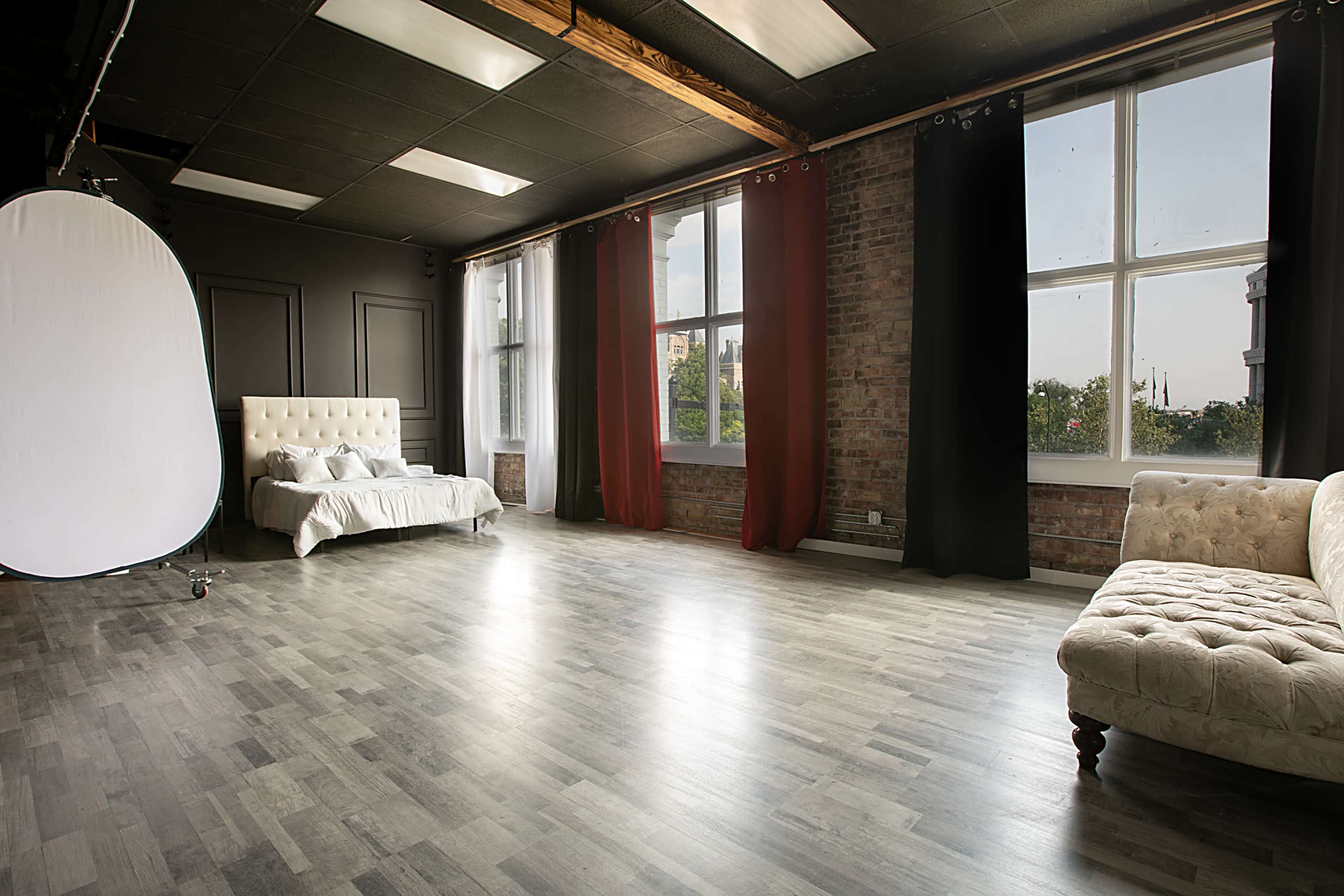 A spacious studio with a white bed, a tufted sofa, large windows with red and black curtains, and a light wooden floor.