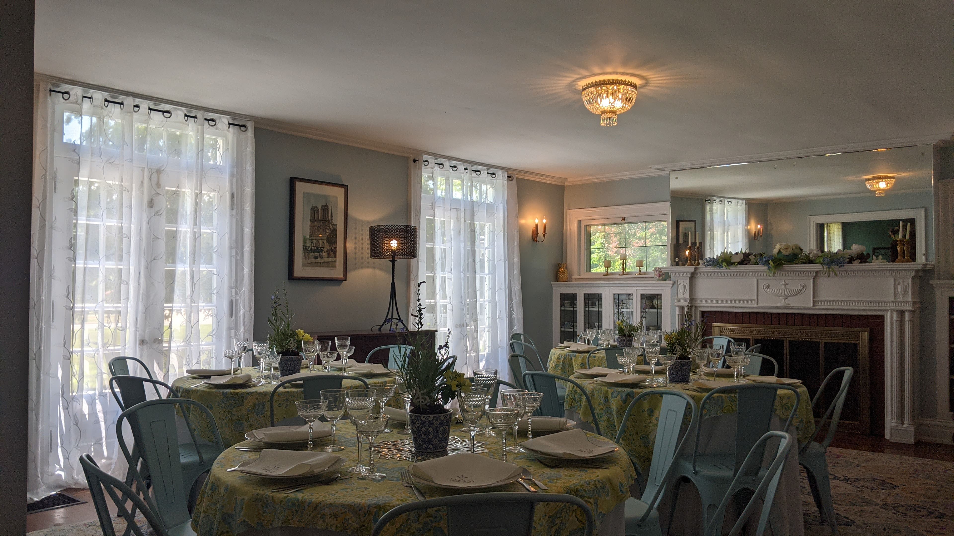 A dining room features a round table set for a meal, surrounded by blue chairs, with large windows and a mirror reflecting the decor.