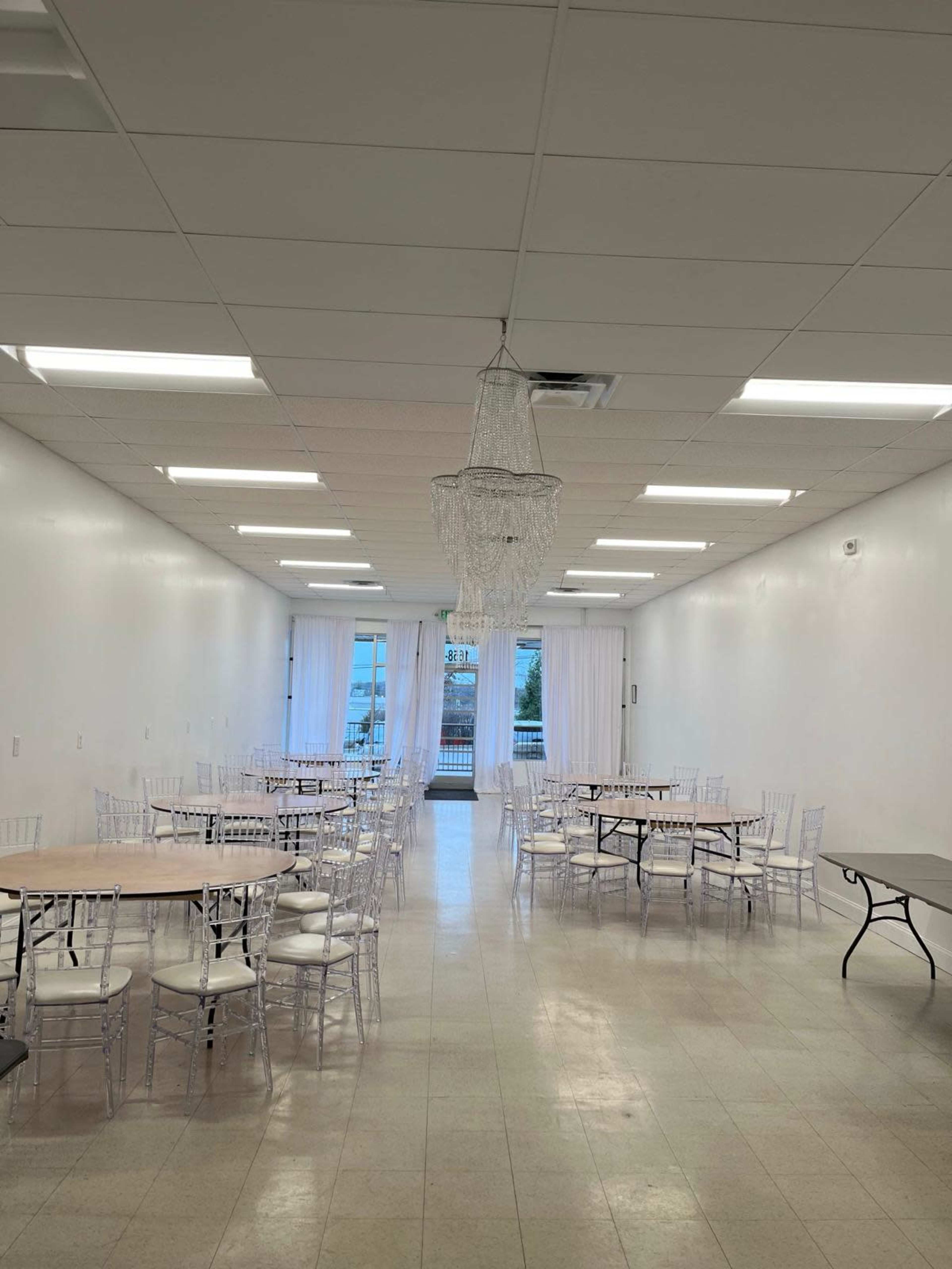 The image shows a spacious, brightly lit room with several round tables and clear chairs arranged neatly, featuring a chandelier hanging from the ceiling.