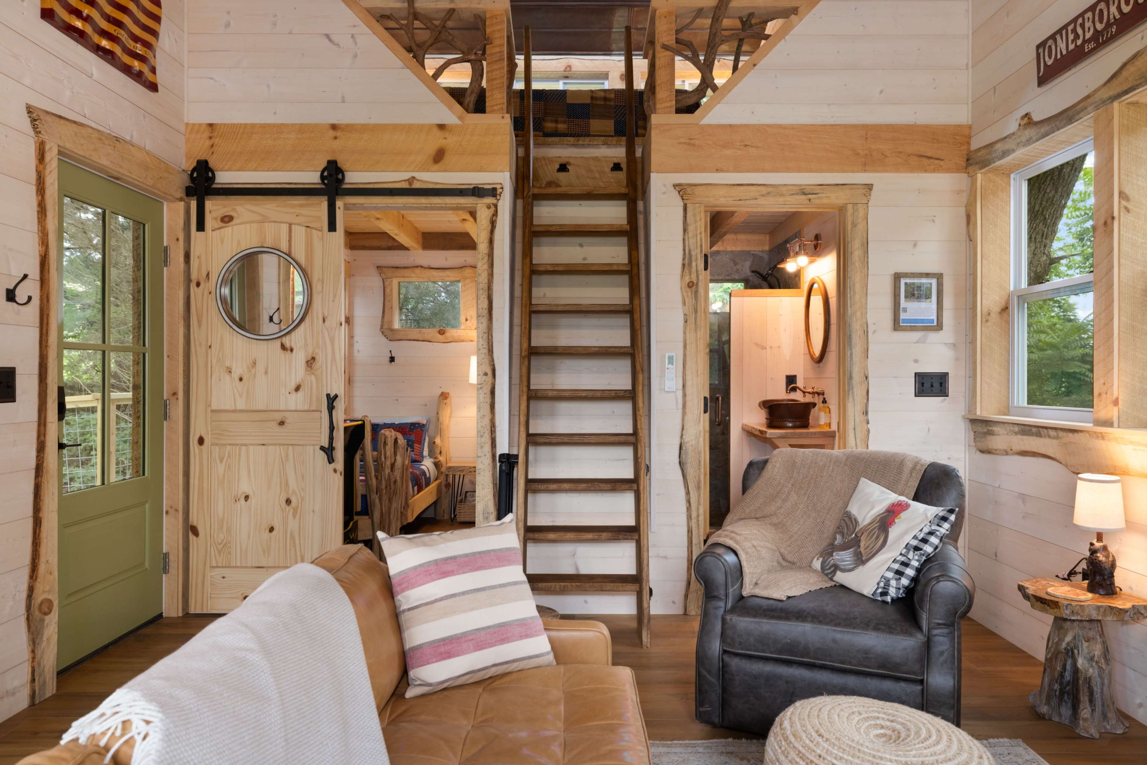 The image shows the interior of a cozy cabin with wooden walls, a staircase leading to an upper level, a living area featuring a sofa and an armchair, and a bathroom visible through a door on the right.