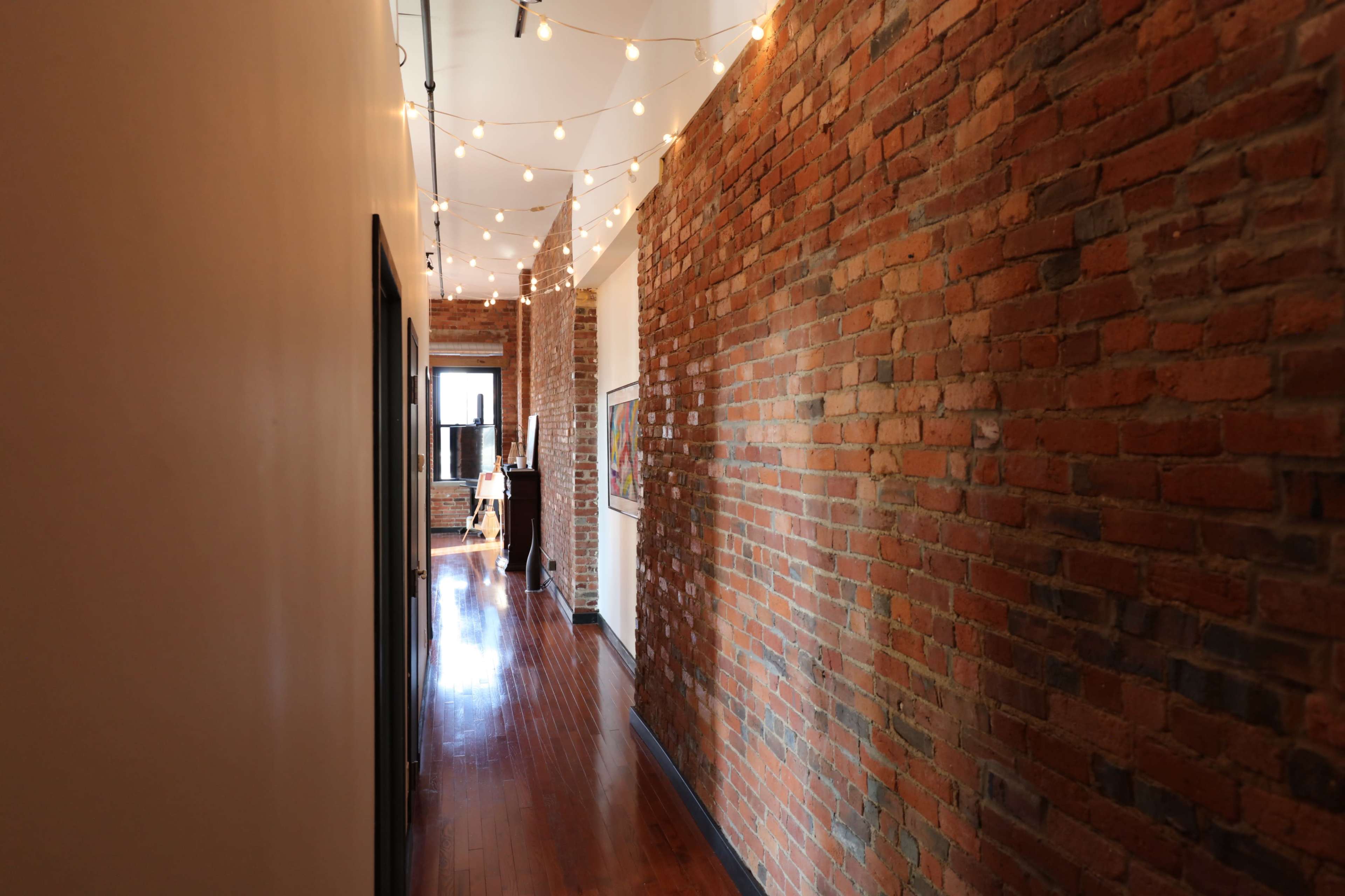 A narrow hallway with exposed brick walls, polished wooden floors, and string lights overhead.