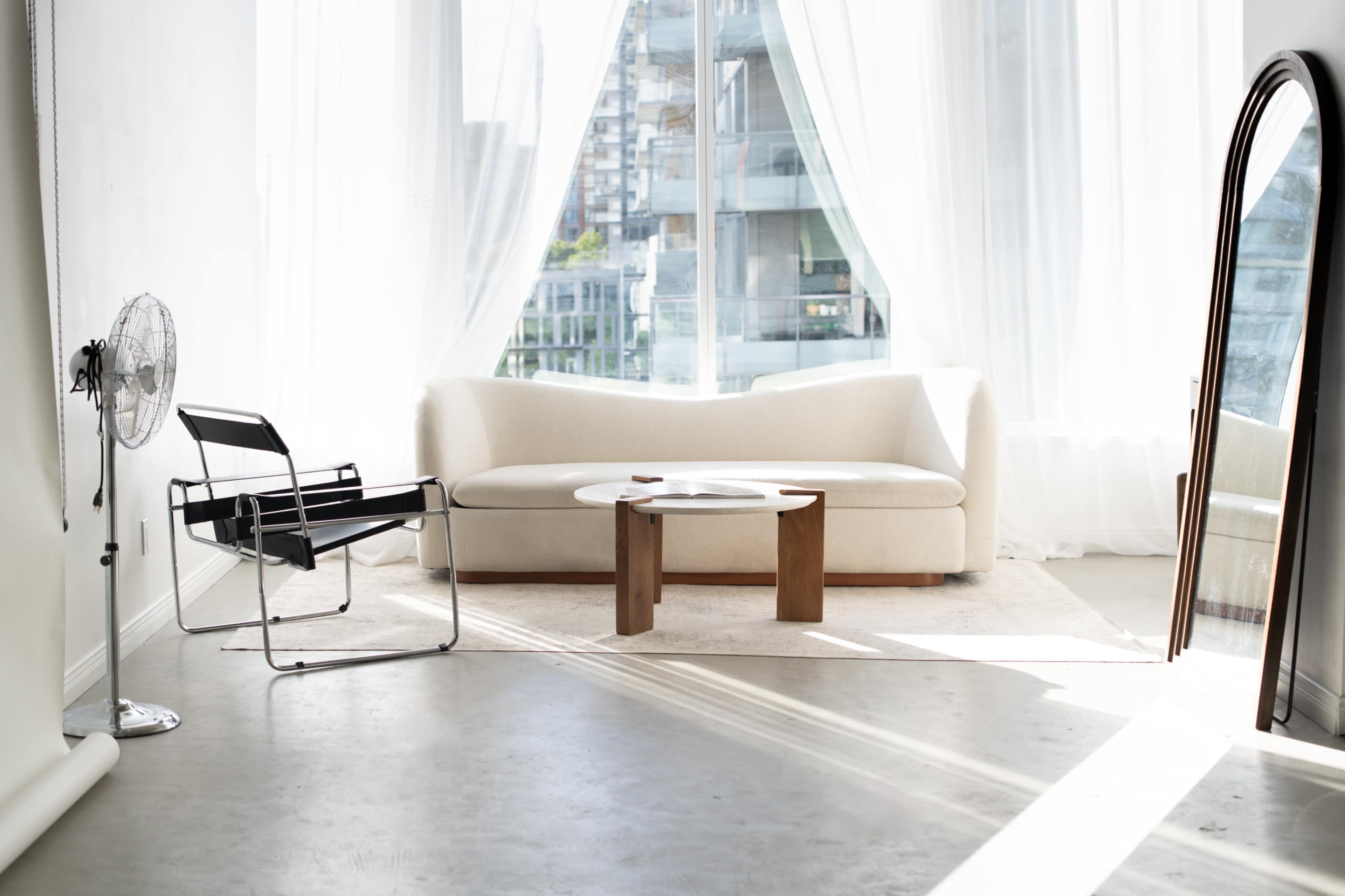 The image shows a modern, minimalist living room with a white sofa, a wooden coffee table, and a chair, illuminated by natural light coming through large windows.