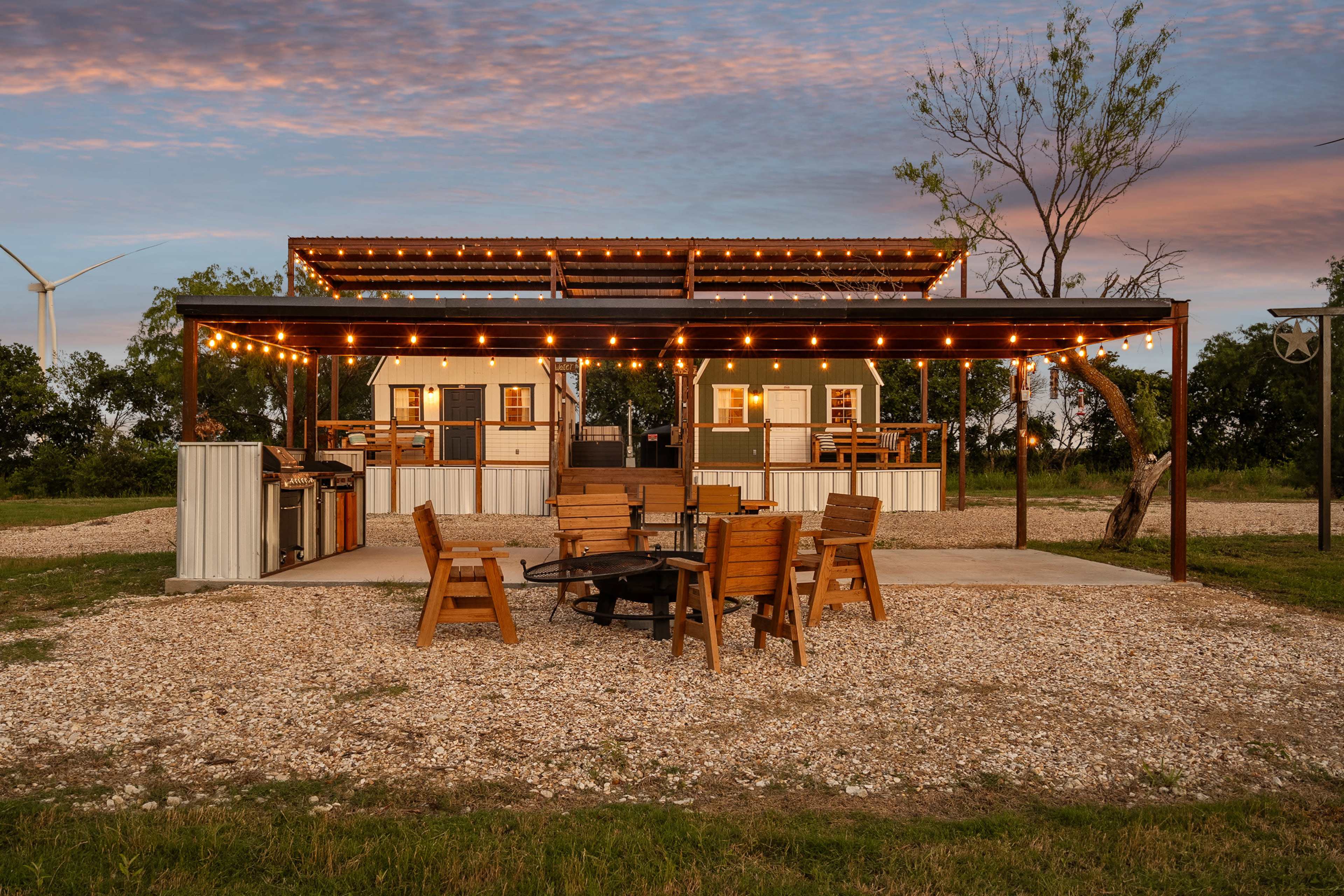 The image shows a modern outdoor seating area with a fire pit, surrounded by gravel, and a structure with string lights in the background.