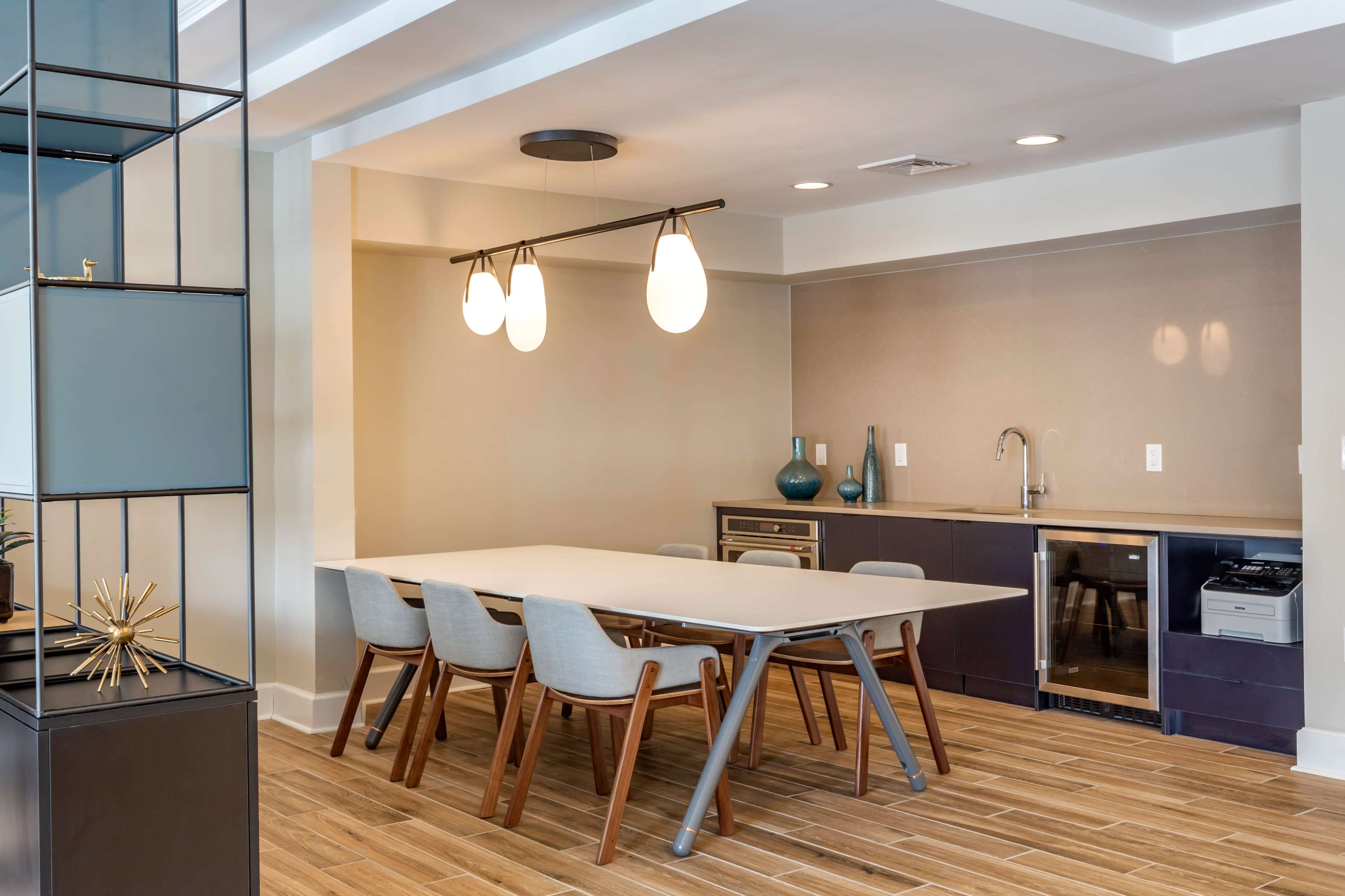 A modern dining area features a long table with wooden legs surrounded by light-colored chairs, adjacent to a kitchenette with dark cabinetry.