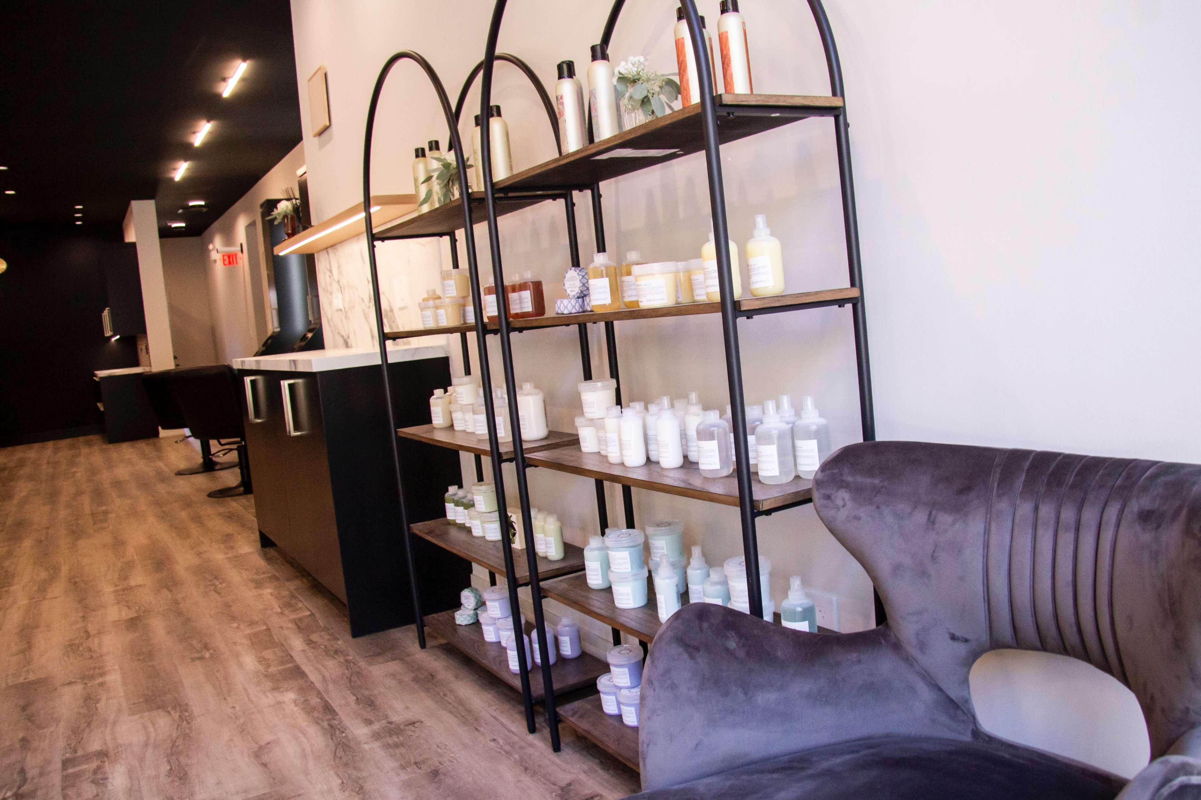 The image shows a modern interior space with a black and white color scheme, featuring a shelving unit displaying various skincare products and a plush gray chair.