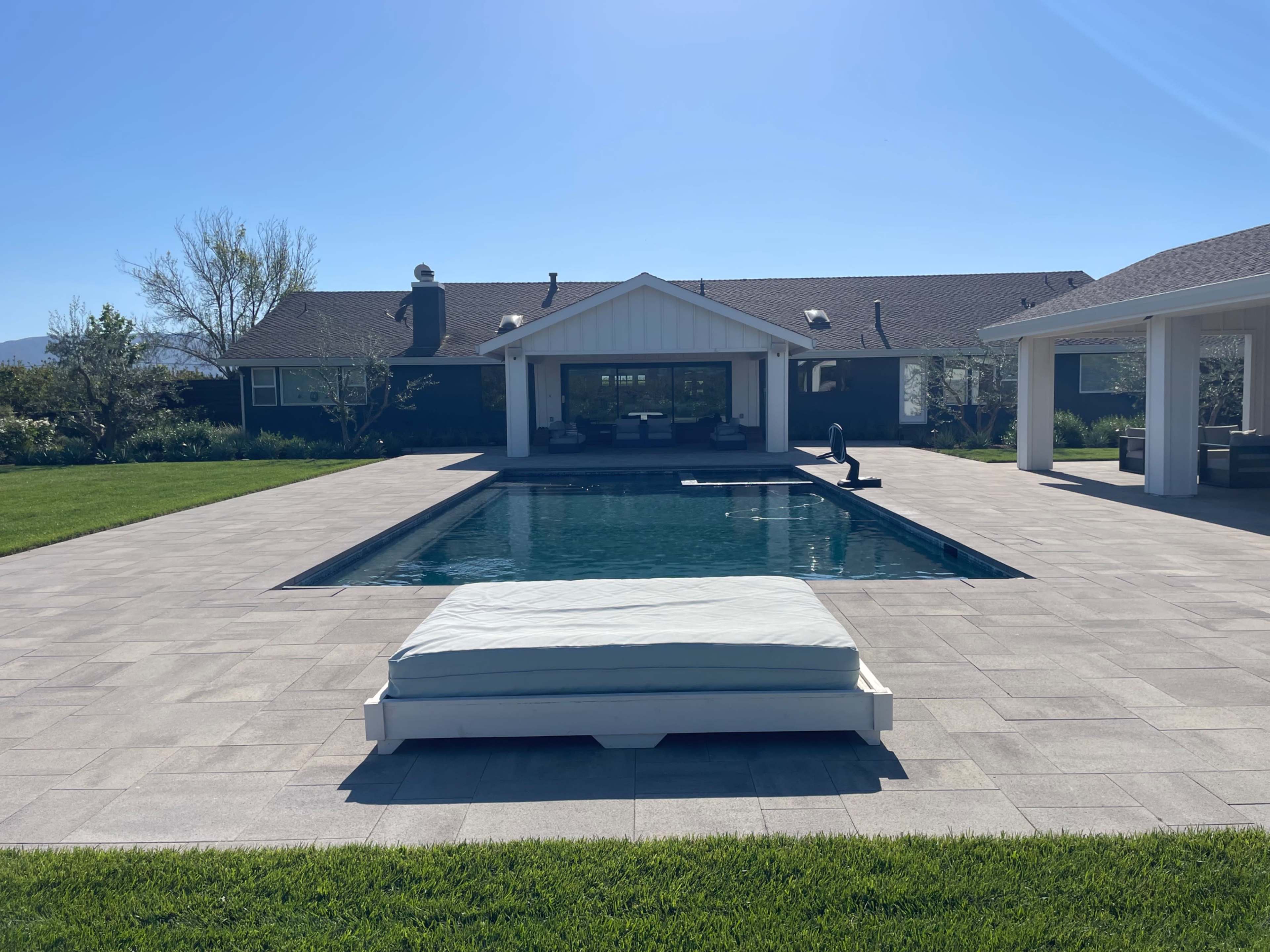 A rectangular swimming pool is surrounded by patio tiles and a grassy area, with a covered seating area visible in the background.