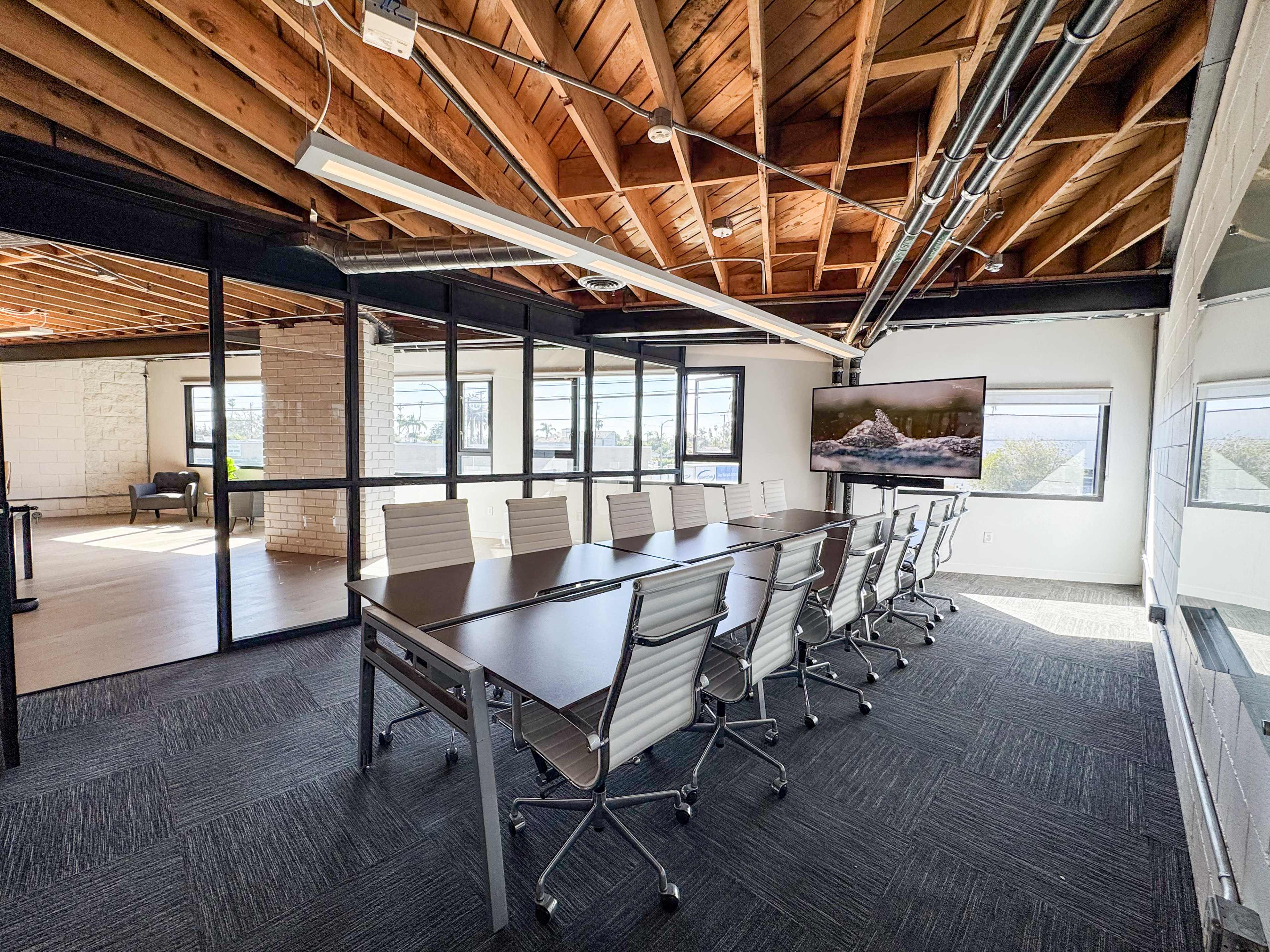 The image shows a modern conference room with a large table, ergonomic chairs, and a TV displaying a nature scene, situated in a well-lit space with exposed wooden beams.