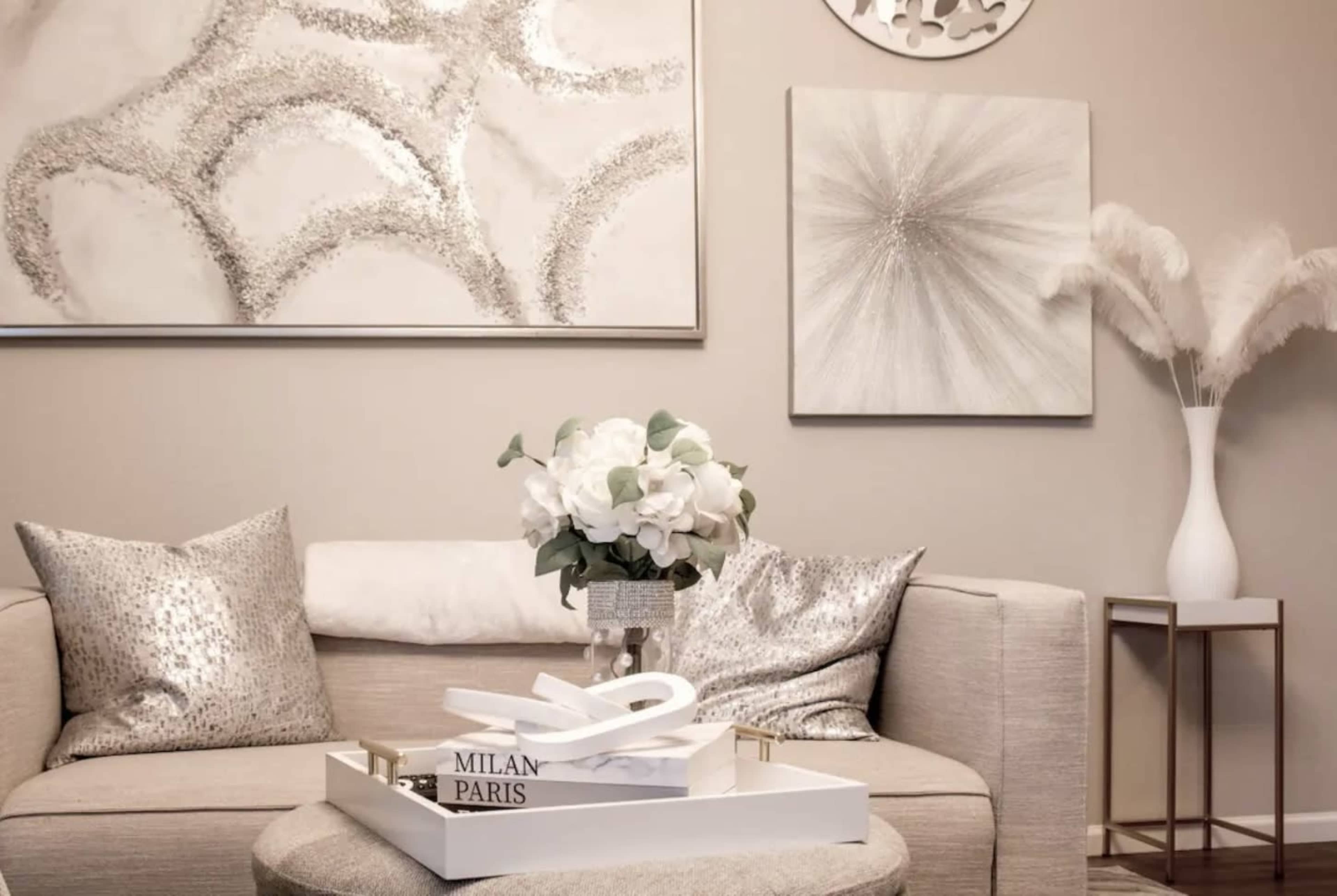 A modern living room features a light-colored sofa adorned with decorative pillows, a white floral arrangement on a central tray, and abstract artwork on the walls.