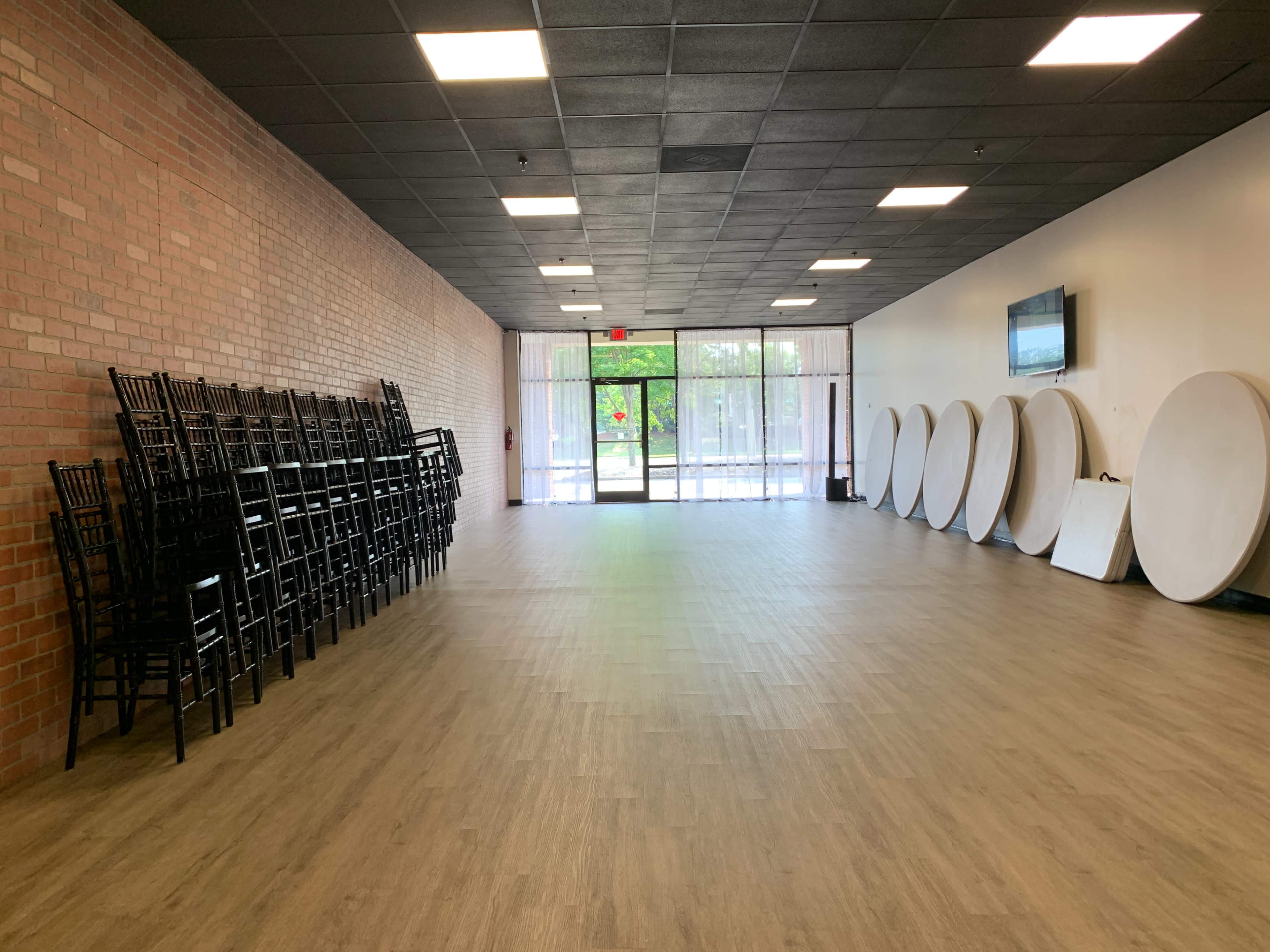 A spacious interior room with stacked chairs against a brick wall, round tables lined up along the opposite side, and large windows at the entrance.