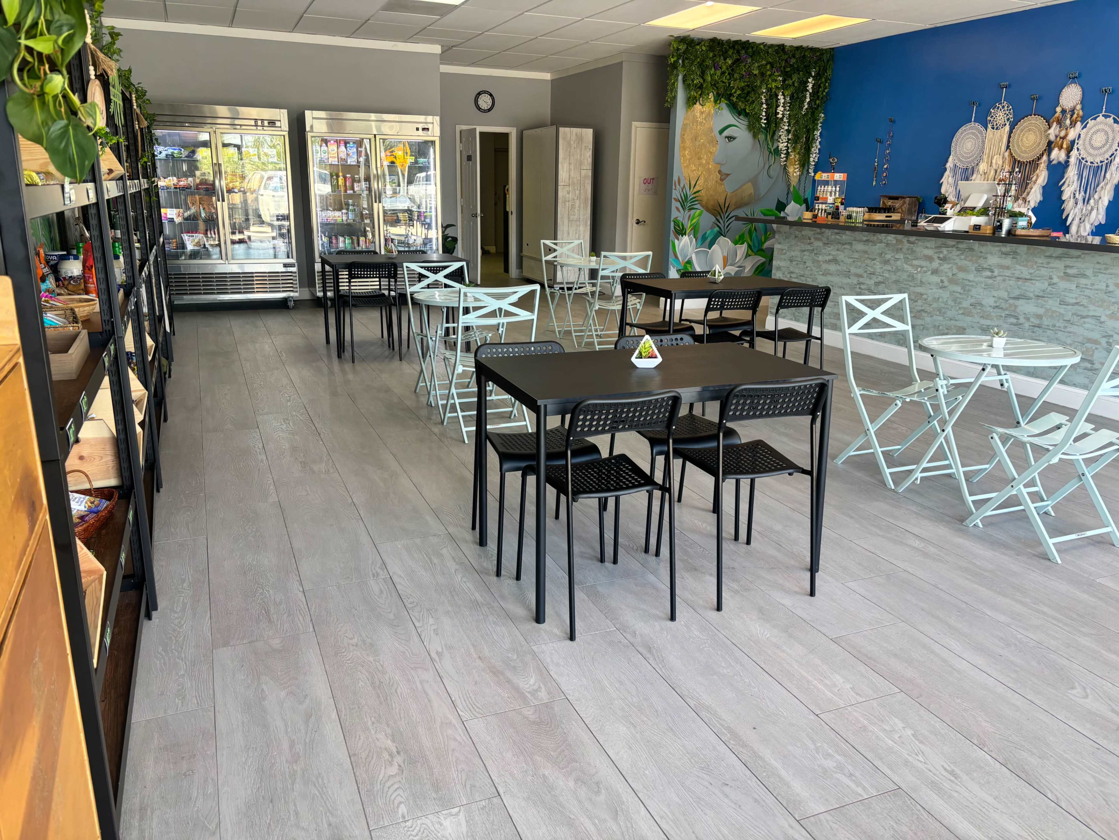The image shows a modern café with tables and chairs arranged inside, featuring a counter and a wall decorated with plants and dreamcatchers.