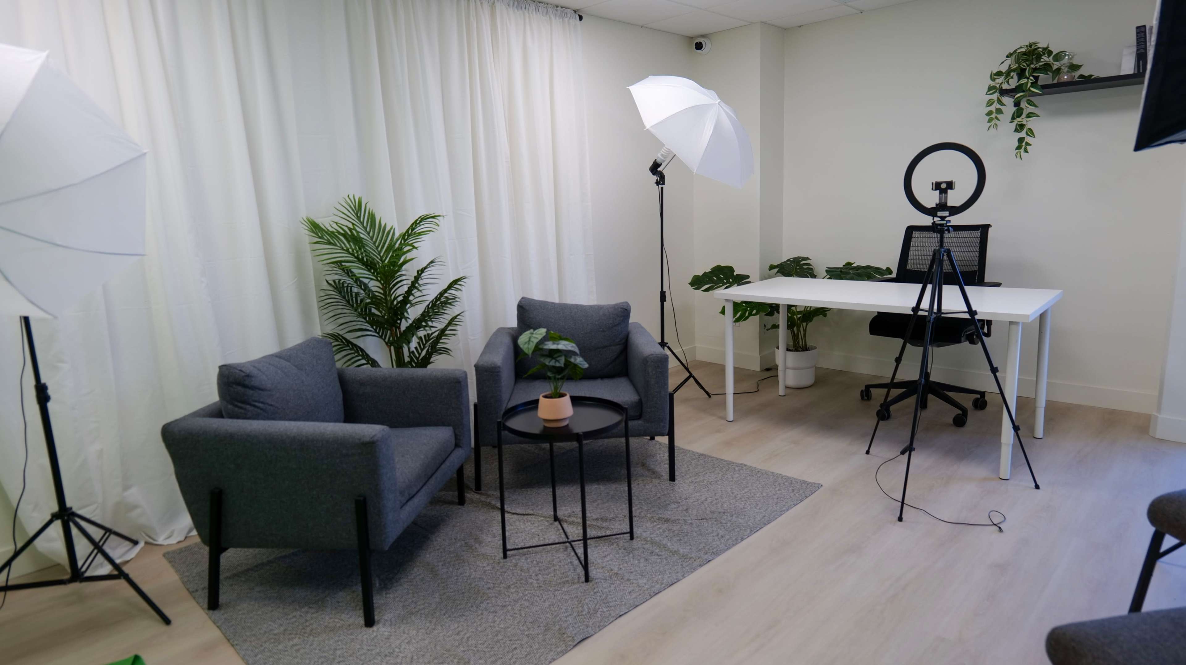 A well-lit, minimally furnished studio space with two gray chairs, a small table with a plant, a photography light setup, and a desk with a ring light.