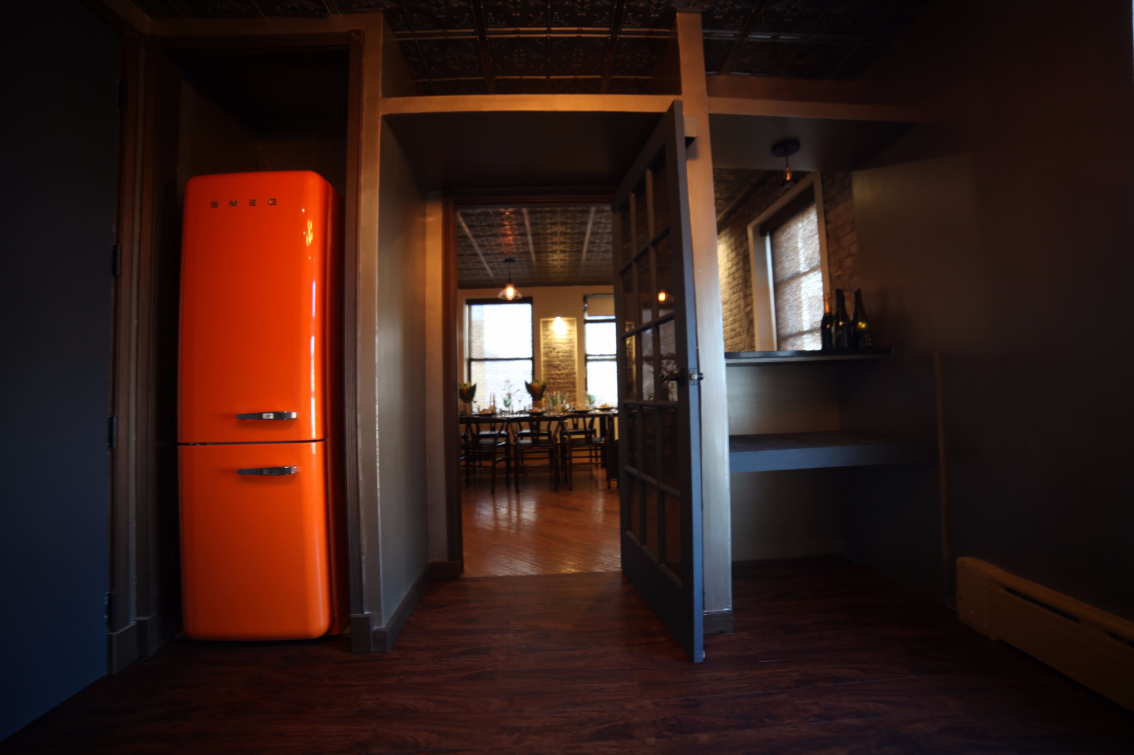 The image shows a brightly colored orange refrigerator beside a doorway leading into a dining area with a wooden table set for a meal.