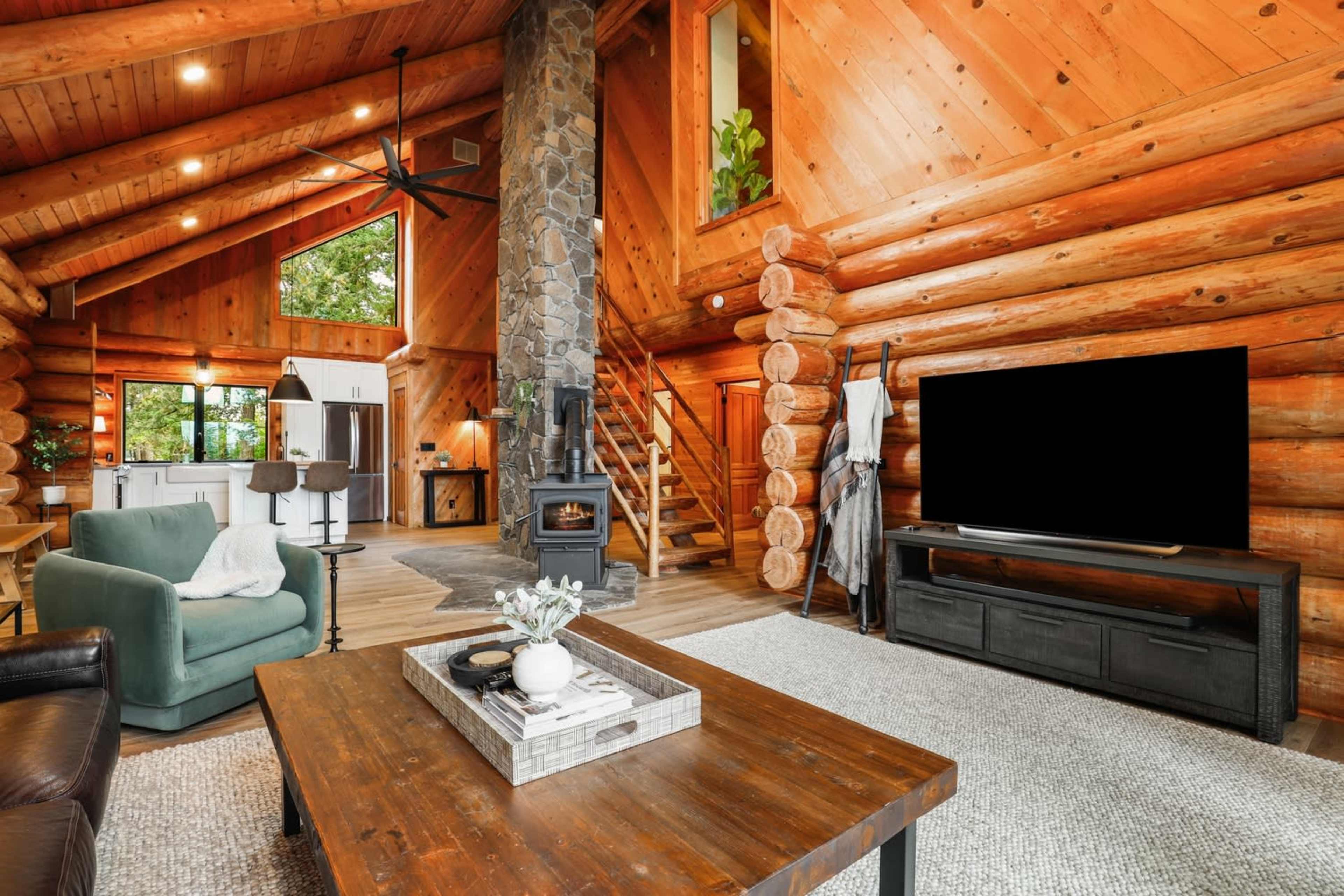 A cozy log cabin interior features a stone fireplace, large windows, and a wooden staircase, with a living area that includes a coffee table and a television.