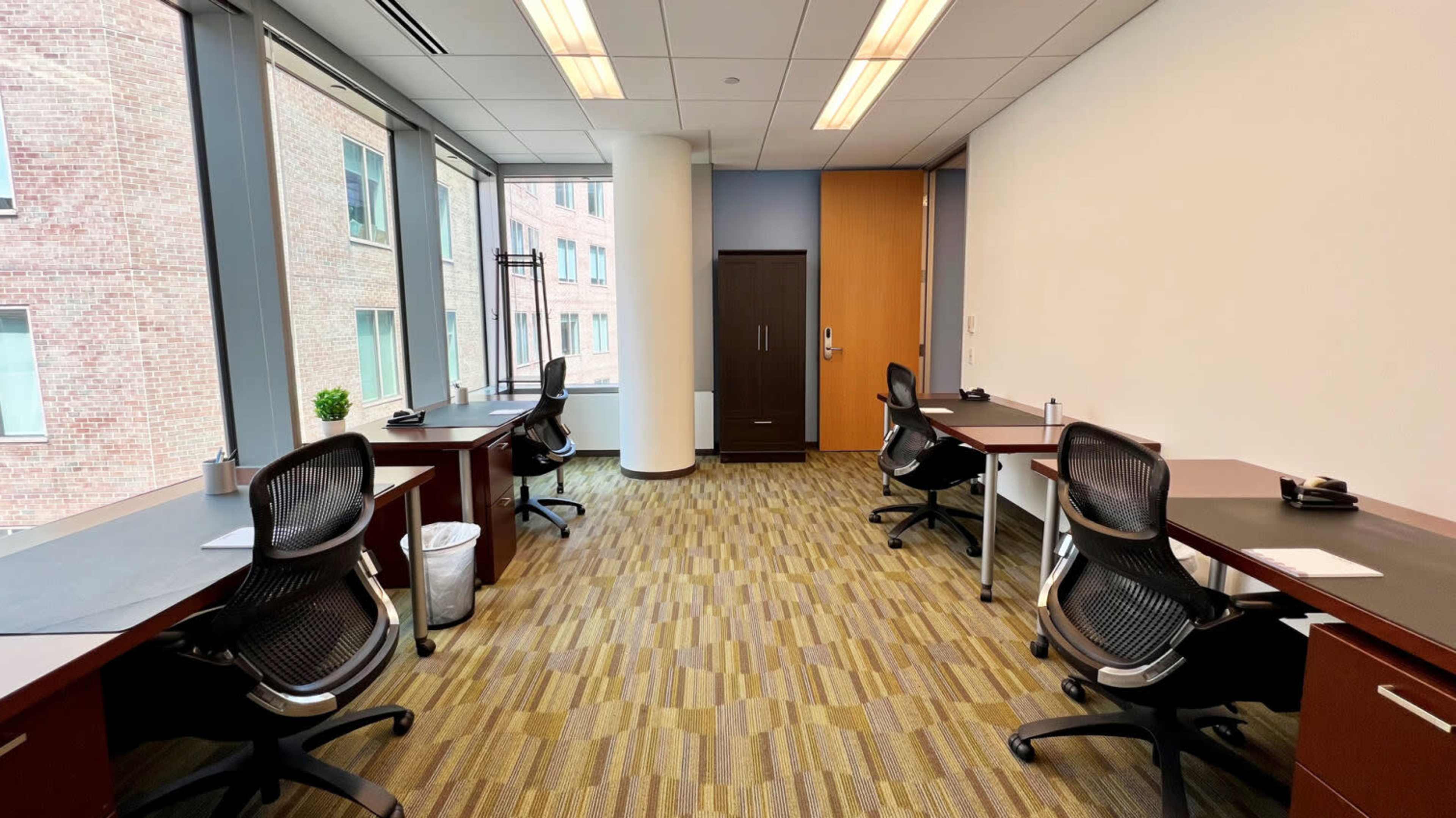 A modern office space with four desks and ergonomic chairs arranged along the walls, featuring large windows letting in natural light.