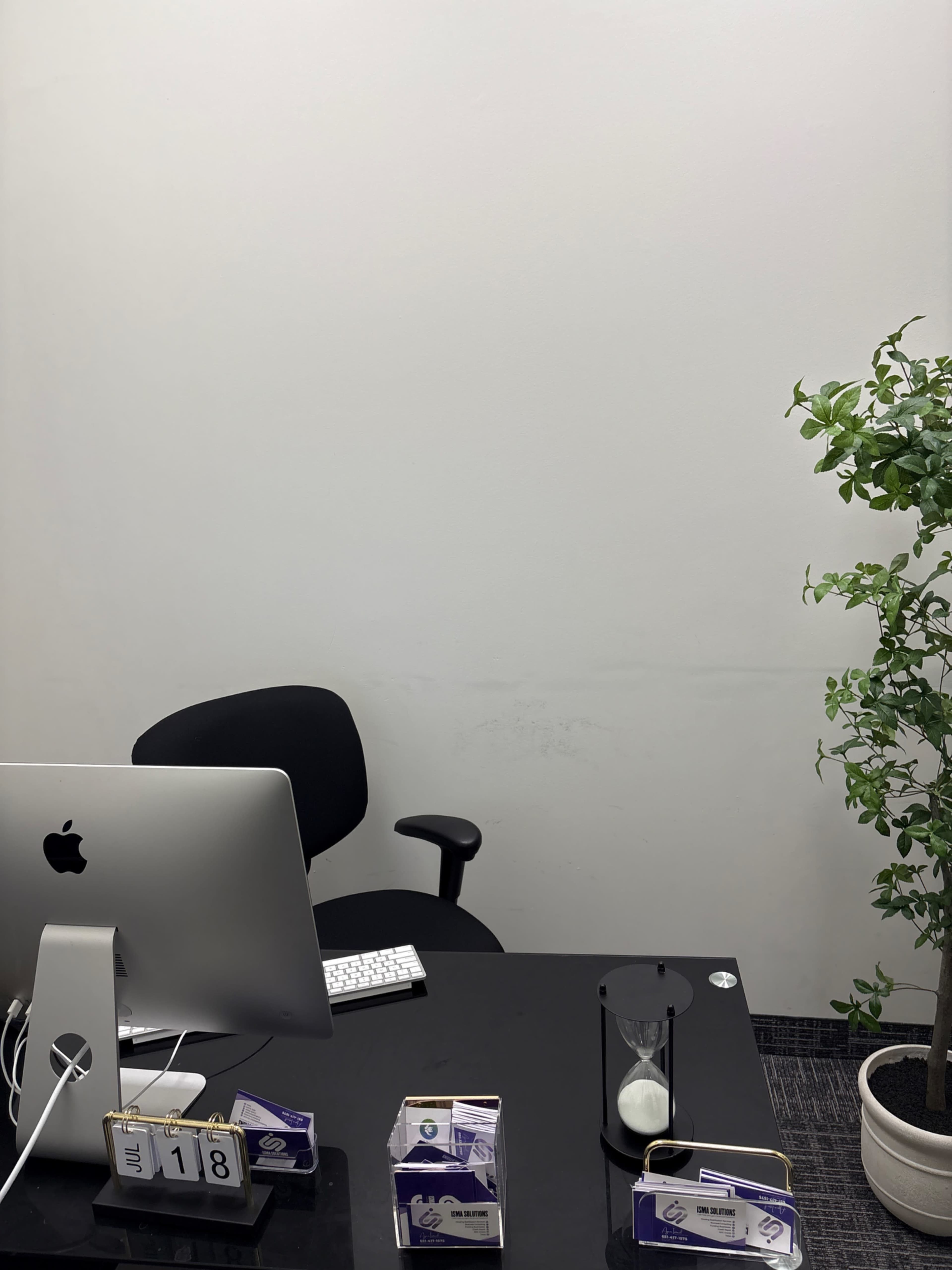 The image shows a minimalist office space with a desk, an office chair, a computer, and a potted plant.