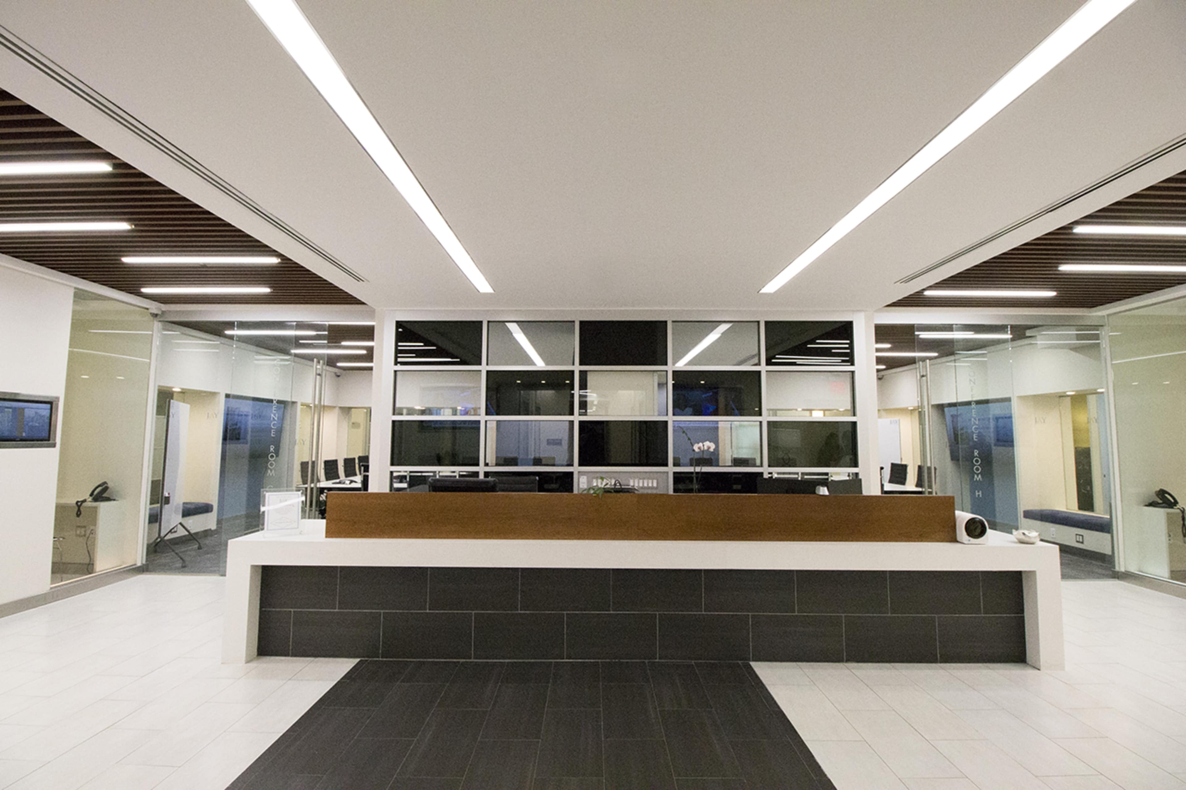 A modern reception area with a central wooden desk and glass partitions, featuring a minimalist design and fluorescent lighting.
