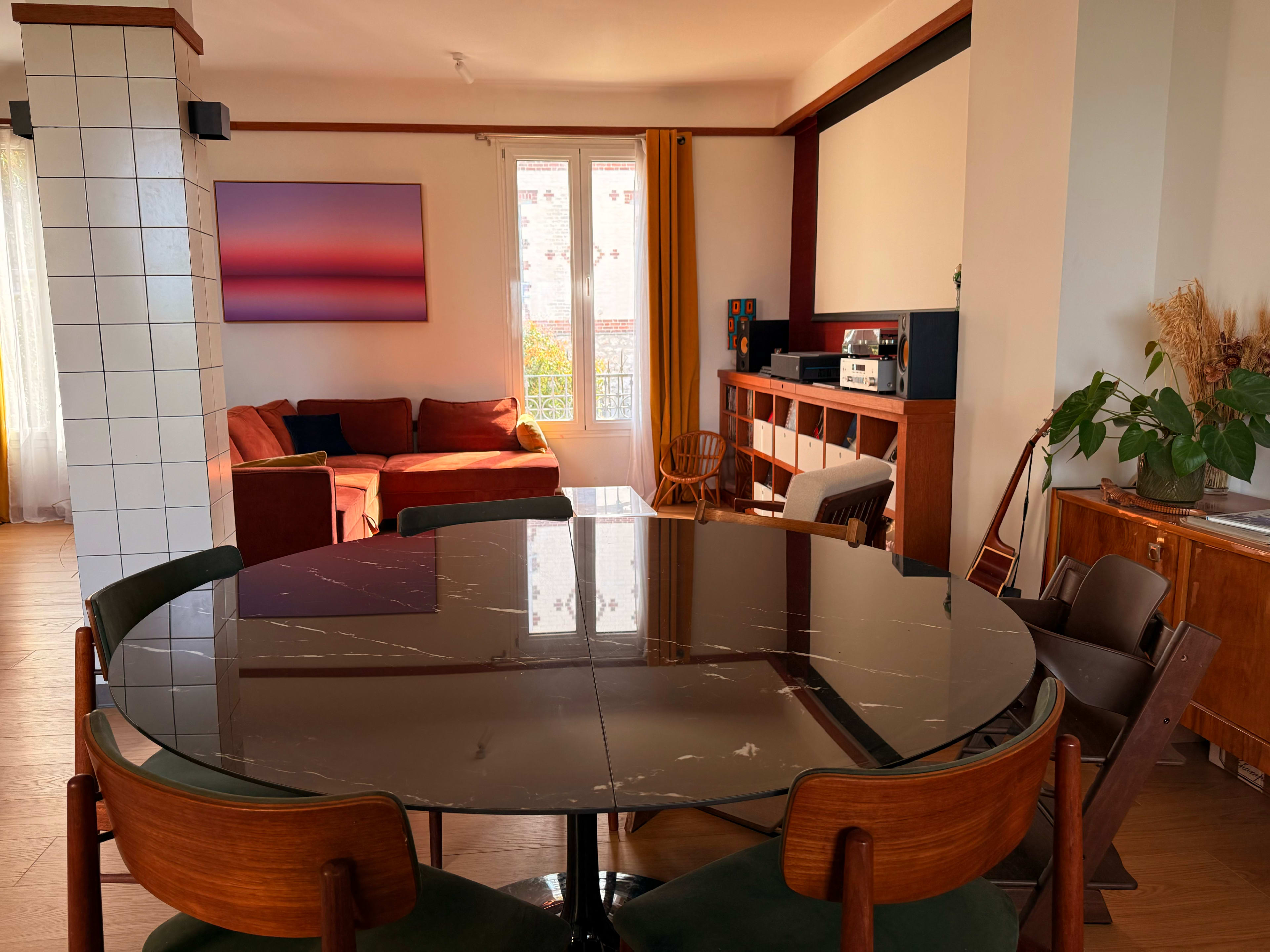 The image shows a modern living space featuring a round dining table, a cozy seating area with a sofa, and large windows allowing natural light to fill the room.