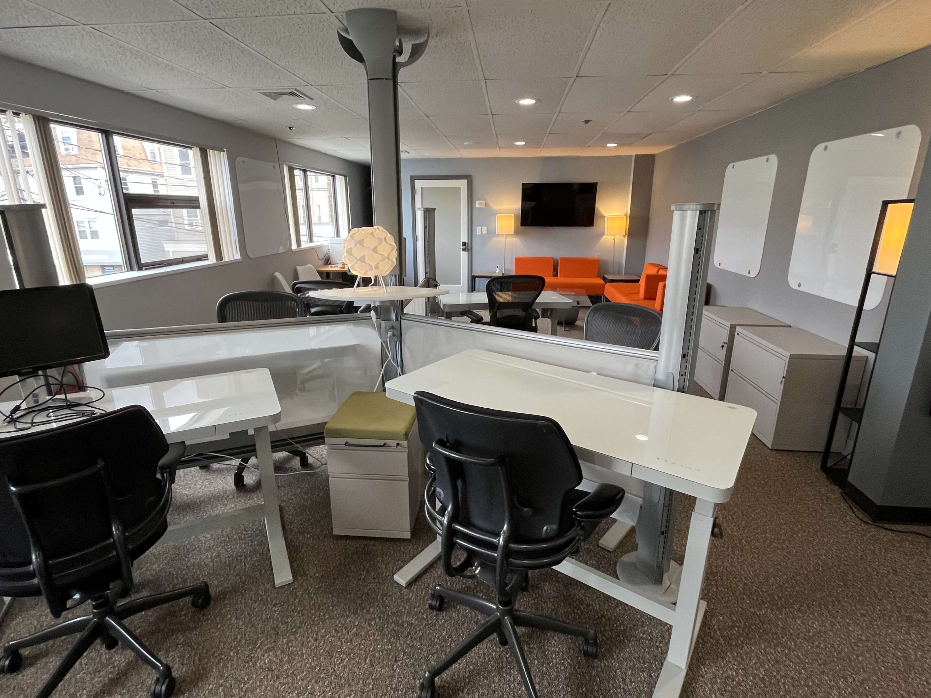 The image shows a modern office space with multiple workstations, a lounge area featuring orange furniture, and large windows allowing natural light.