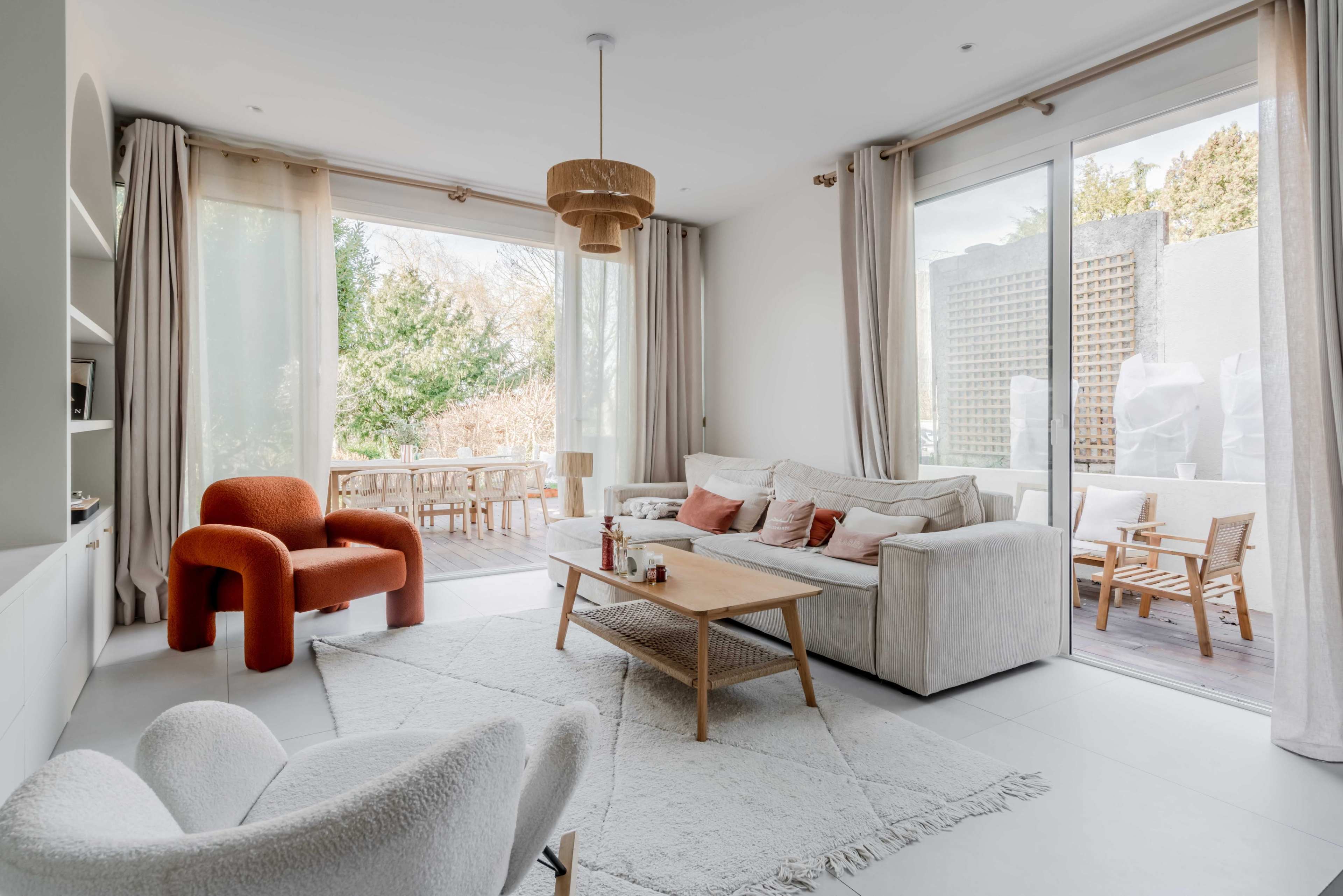 A modern living room with a white sofa, a wooden coffee table, and a red chair, featuring large windows that open to an outdoor space.