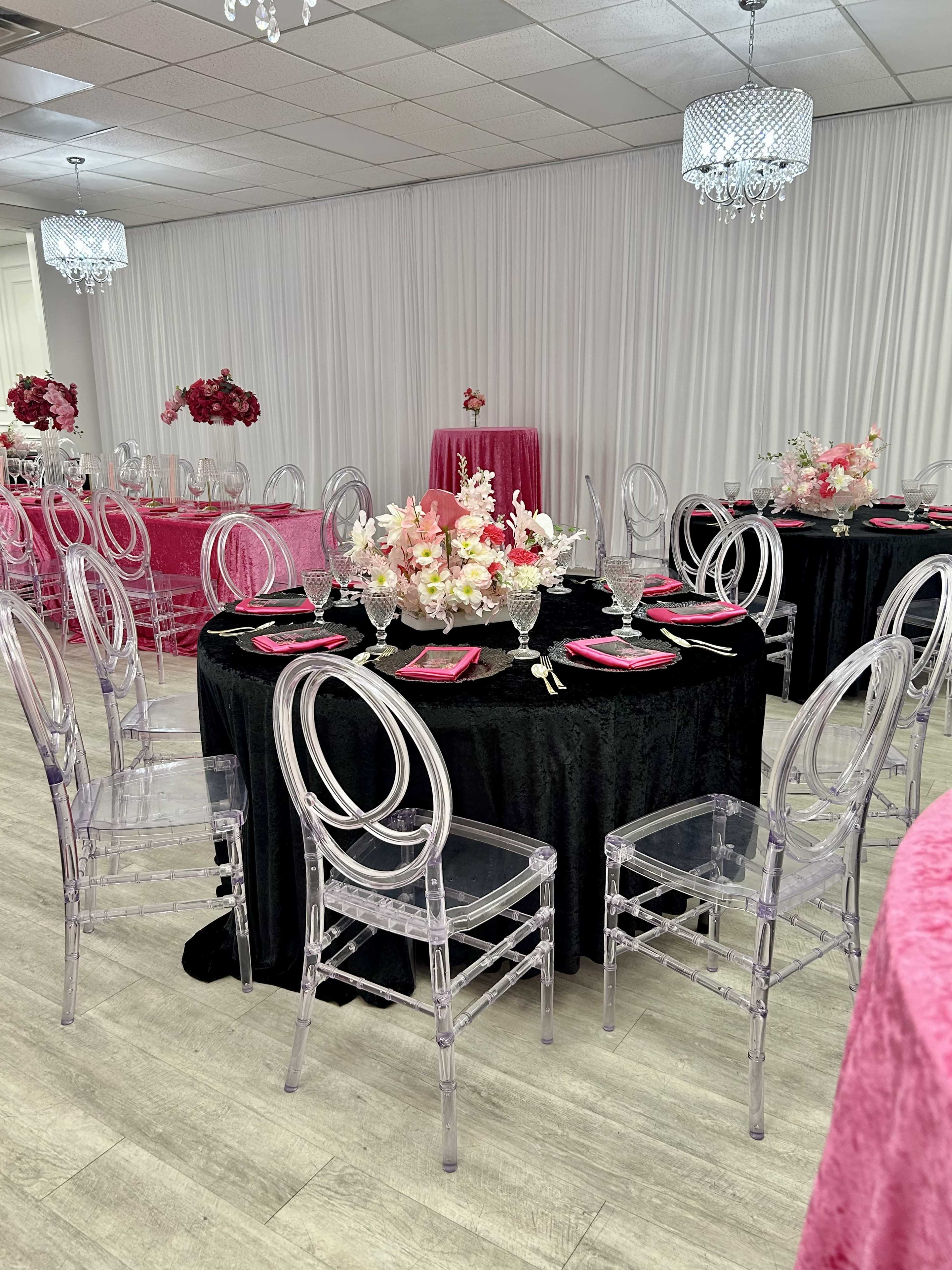 The image shows a decorated event space with tables set in black and pink, featuring clear acrylic chairs and floral centerpieces.