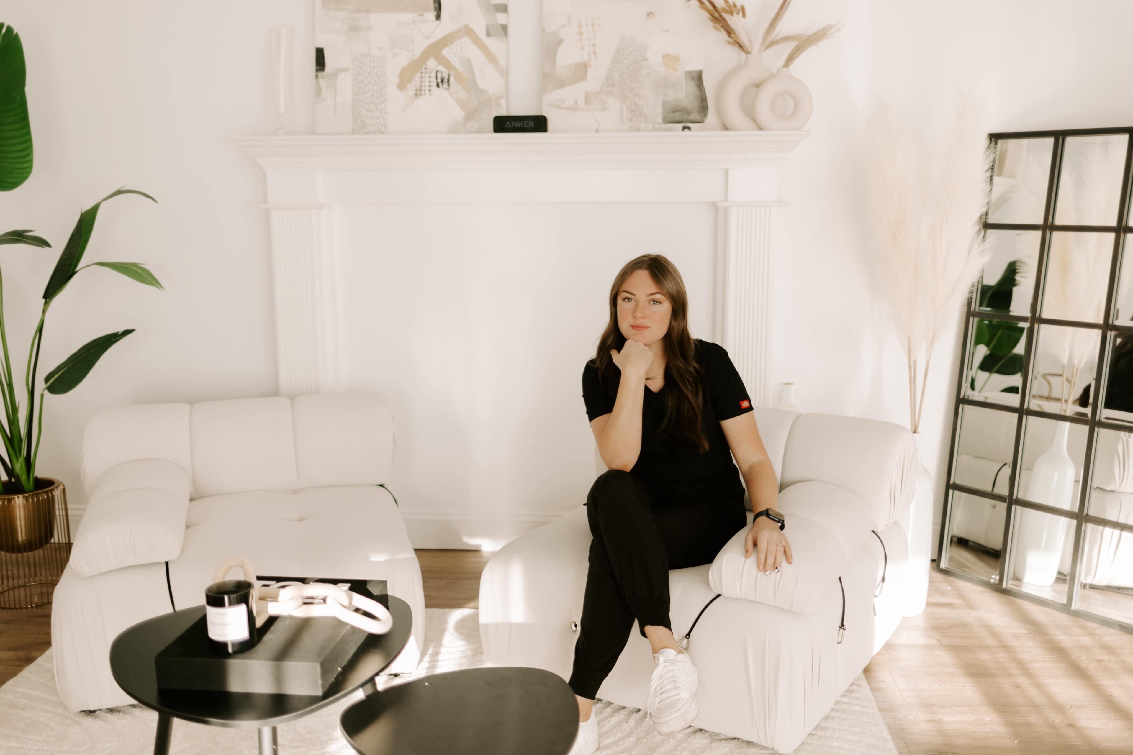 A woman sits thoughtfully on a modern white chair in a bright, minimalist living room.