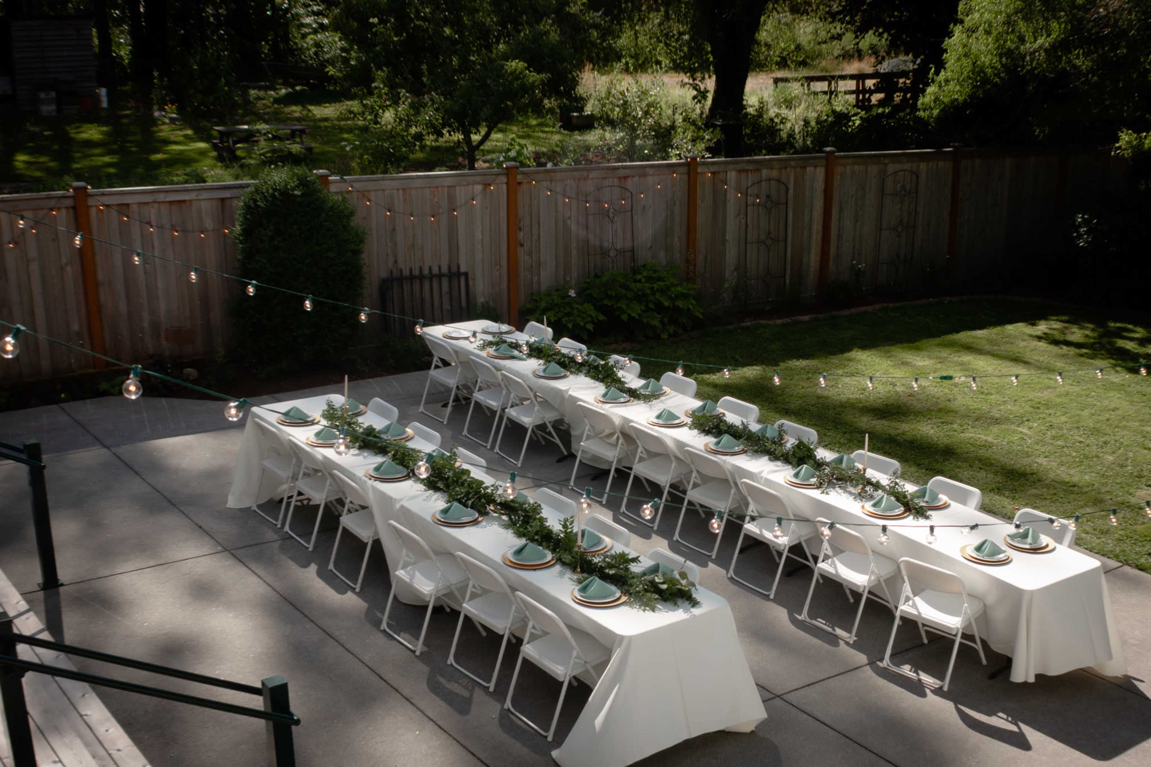 A garden setup with several long tables arranged in a row, each adorned with green table settings and surrounded by string lights.