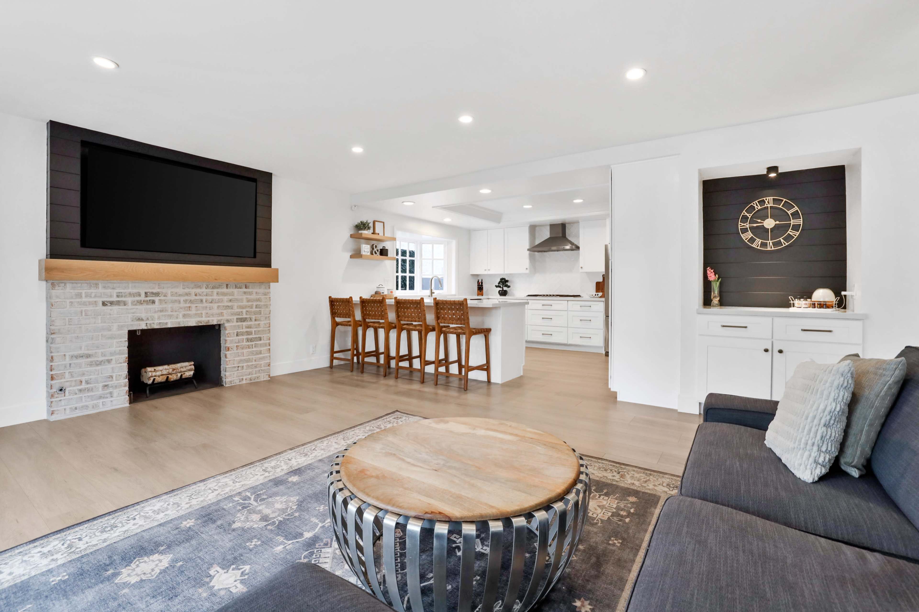 A modern living room features a brick fireplace, a large flat-screen TV mounted above a wooden mantel, and a circular coffee table surrounded by a dark sofa.
