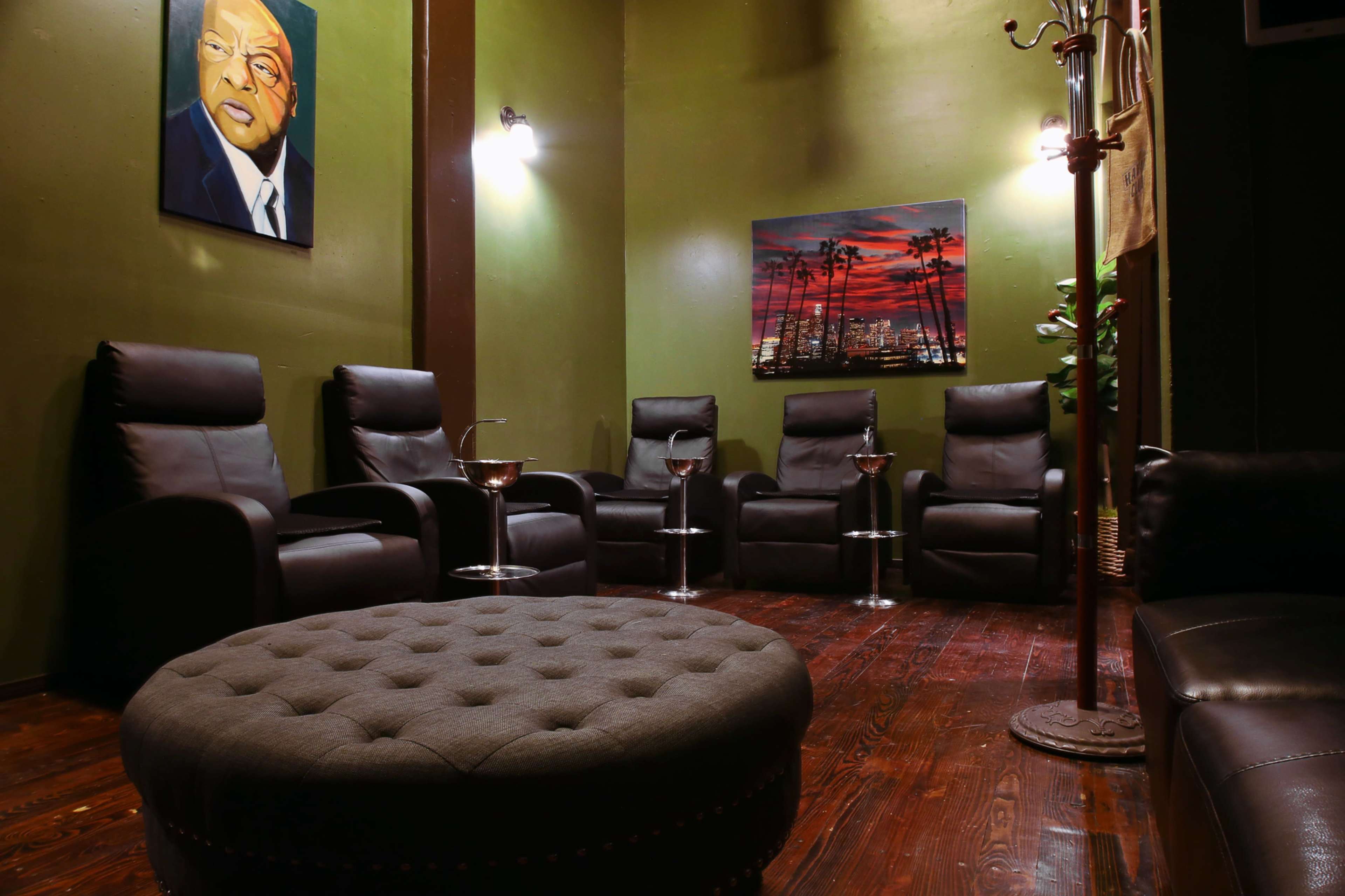 A dimly lit lounge area with five black leather recliners arranged around a round ottoman, featuring two paintings on the walls and small tables with drinks.
