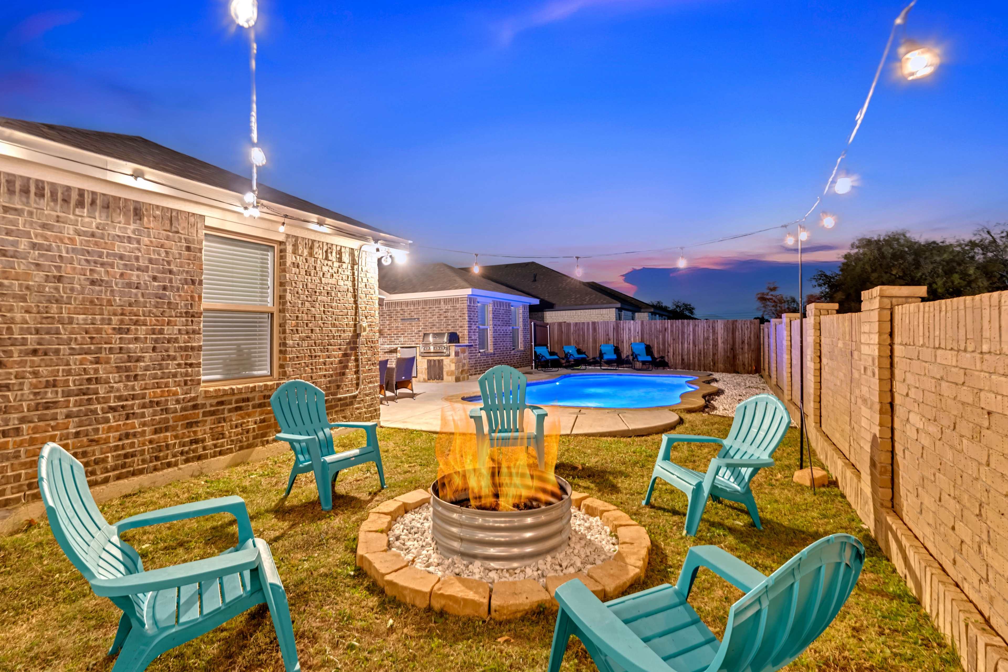 A fire pit surrounded by blue Adirondack chairs sits in a backyard with a pool and string lights strung overhead at dusk.