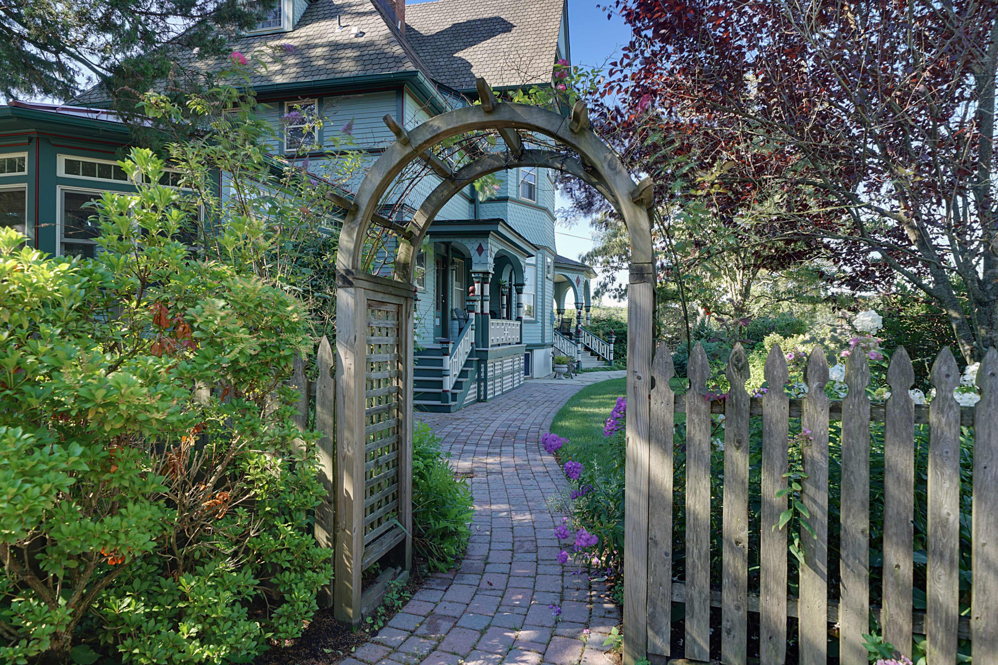 A wooden archway leads to a path of brick pavers bordered by flowers, leading to a house with a green exterior and surrounding gardens.