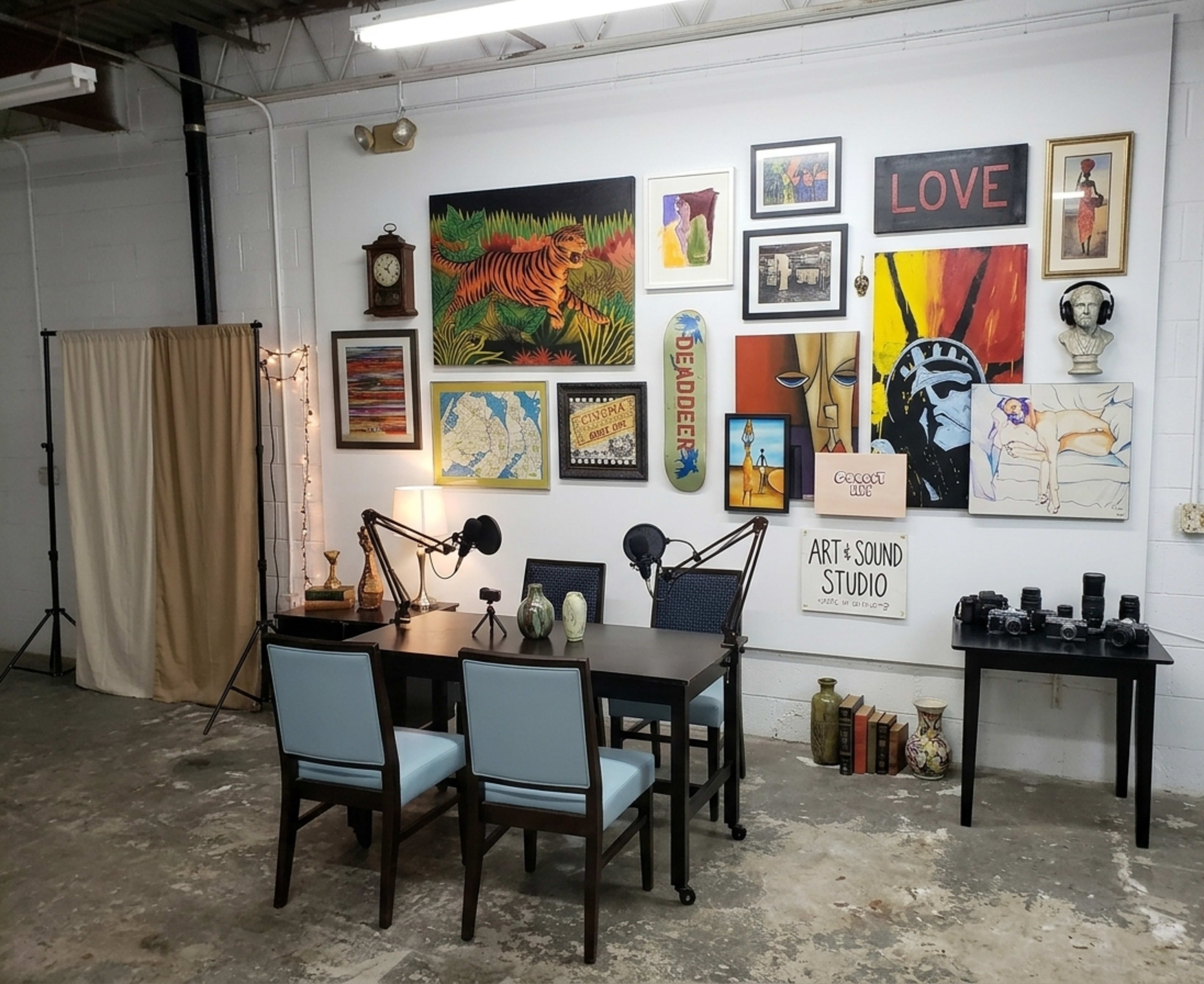 The image shows a studio space featuring a table with four chairs, two microphones, and a gallery wall adorned with various framed artworks and decorations.