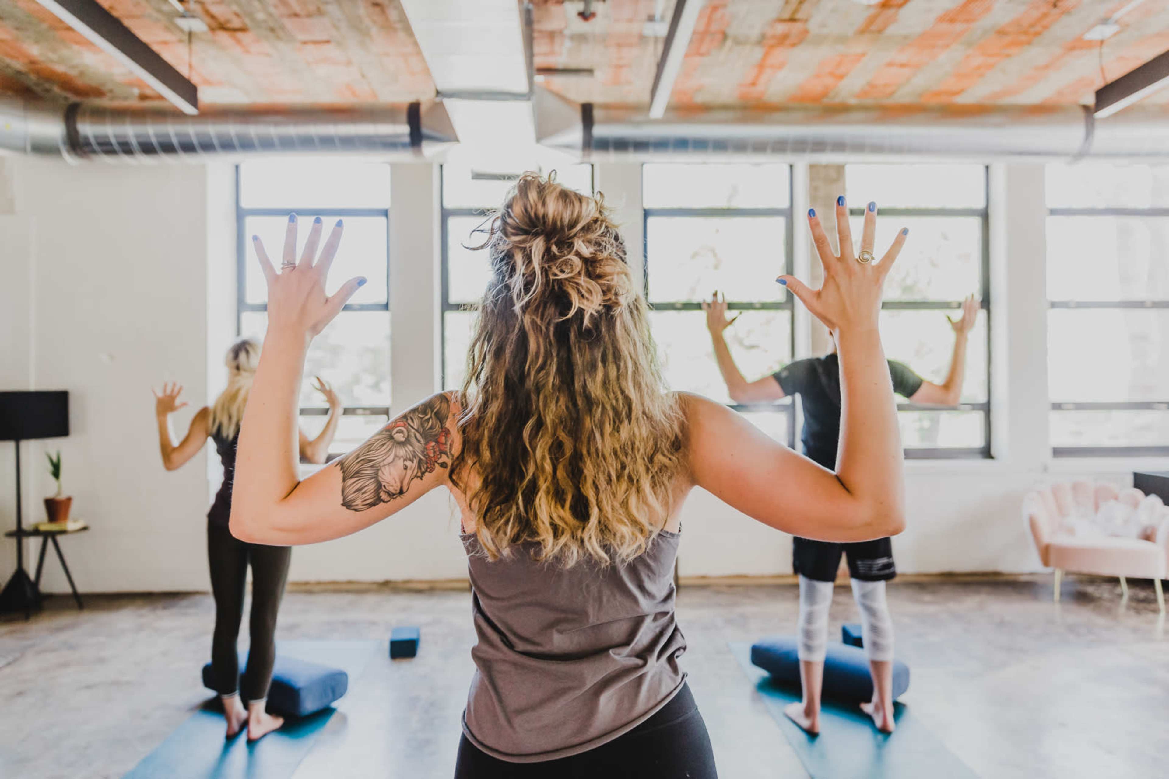 A group of three people is practicing yoga in a well-lit studio, with their backs facing the camera, each raising their arms while standing on yoga mats.