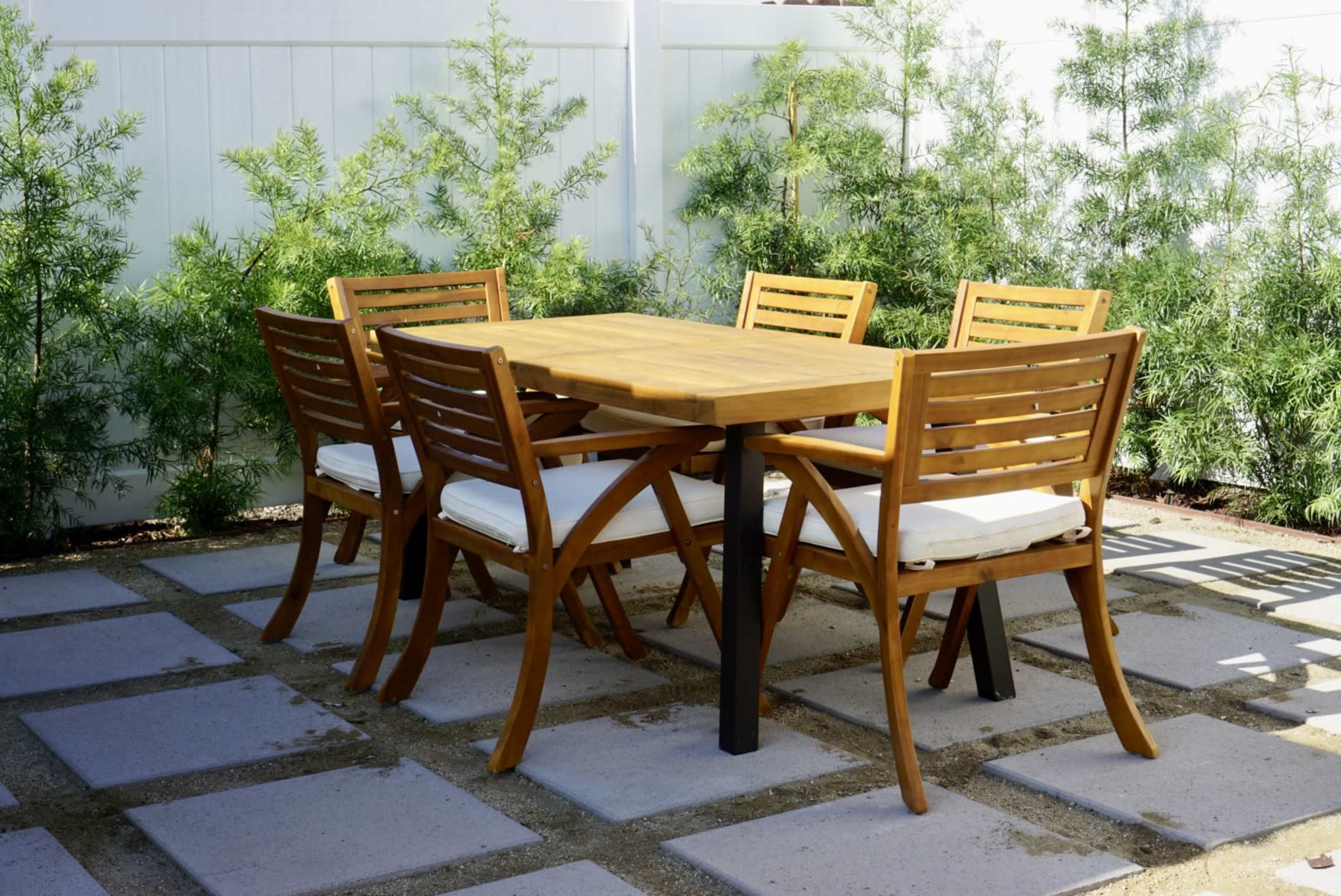 A wooden dining table with six chairs is set on a patio surrounded by small trees.