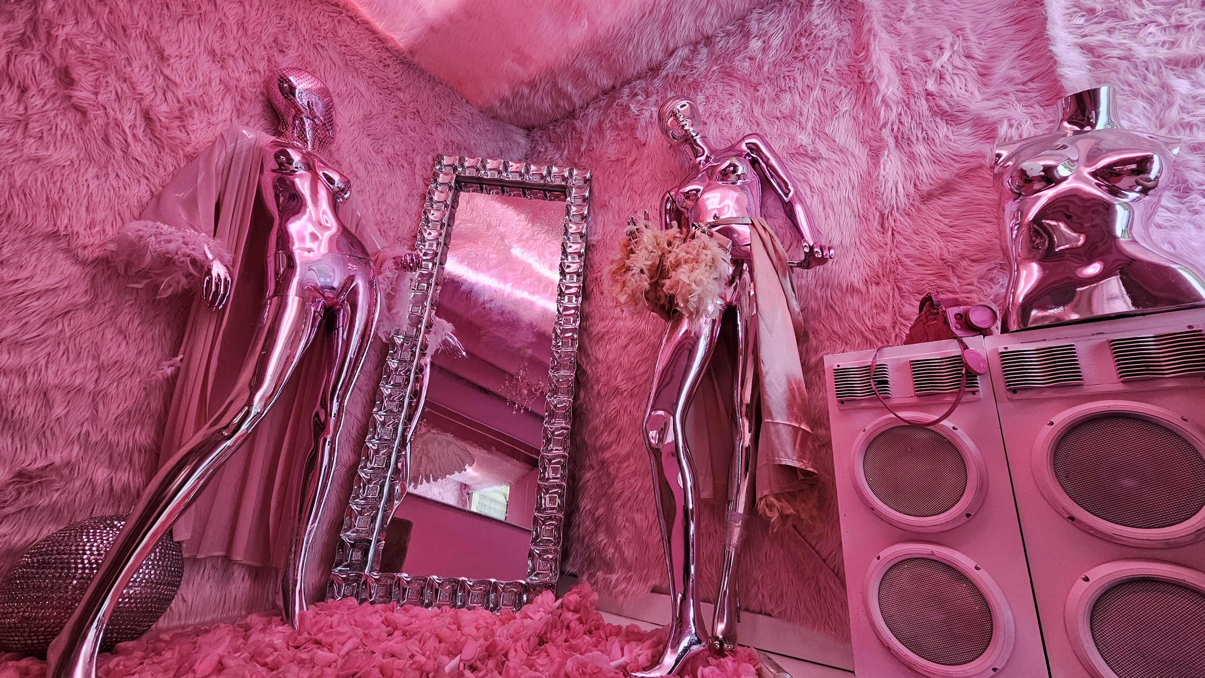 The scene features shiny metallic mannequins in a pink, fur-covered room with a large mirror and vintage speakers.