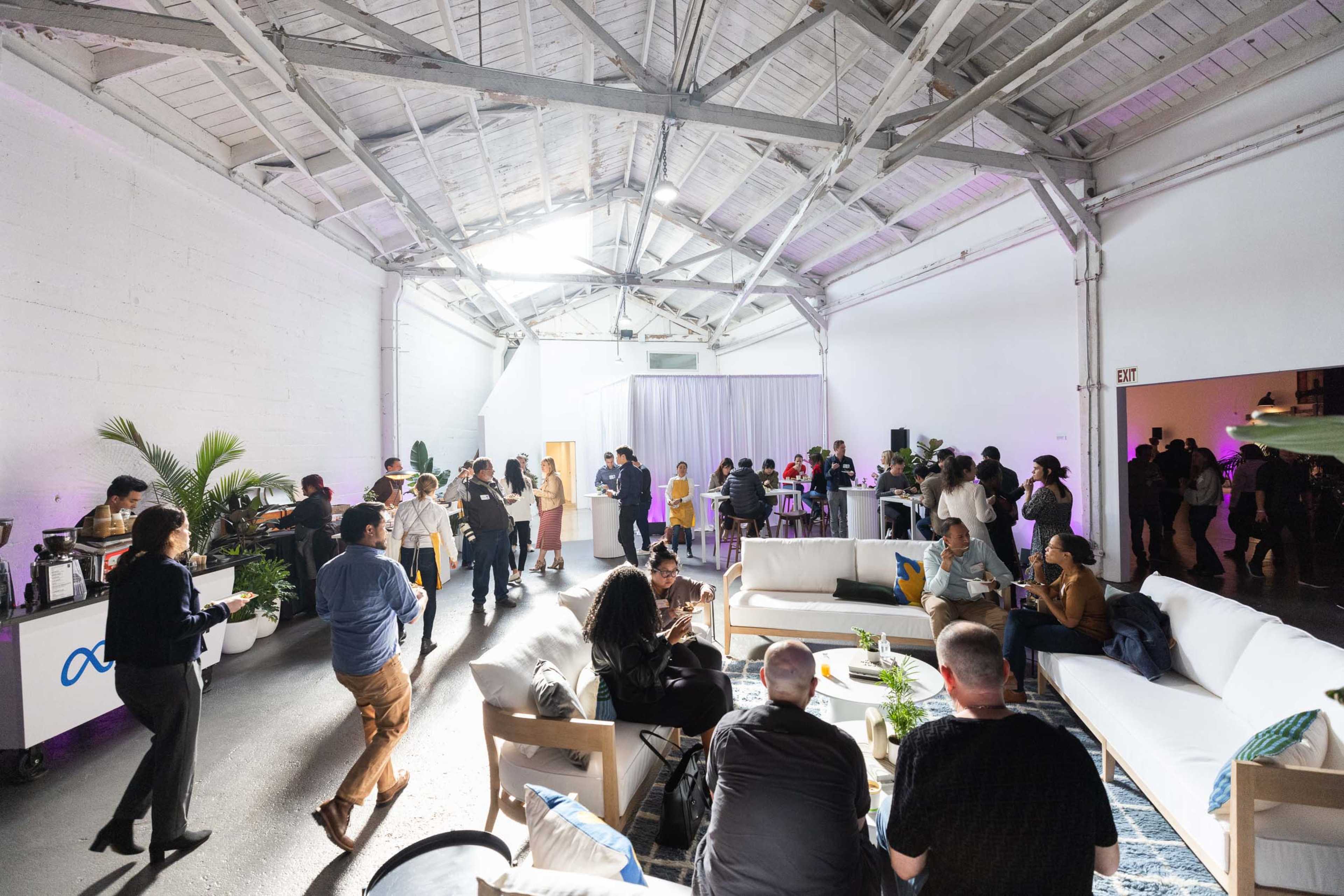 The image shows a lively indoor event with attendees socializing and sitting on couches in a well-lit industrial space.