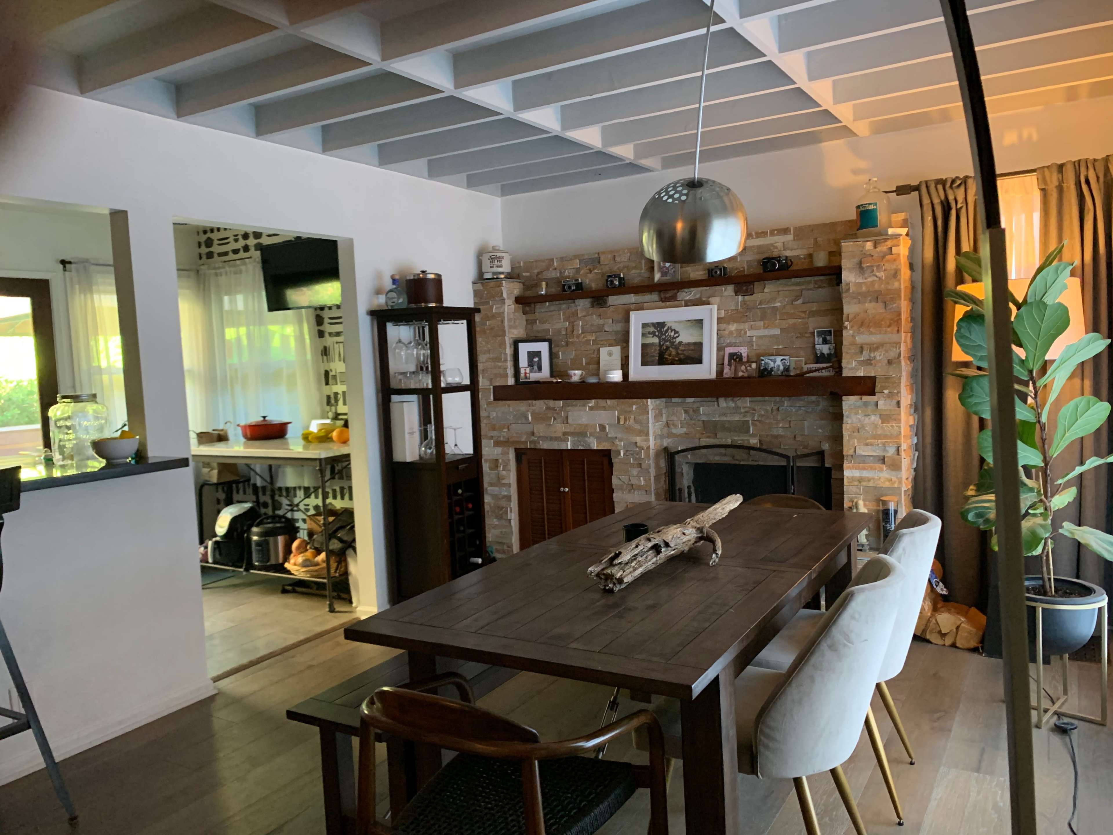 The image shows a dining area with a wooden table, surrounded by chairs, next to a stone fireplace, and an open kitchen in the background.