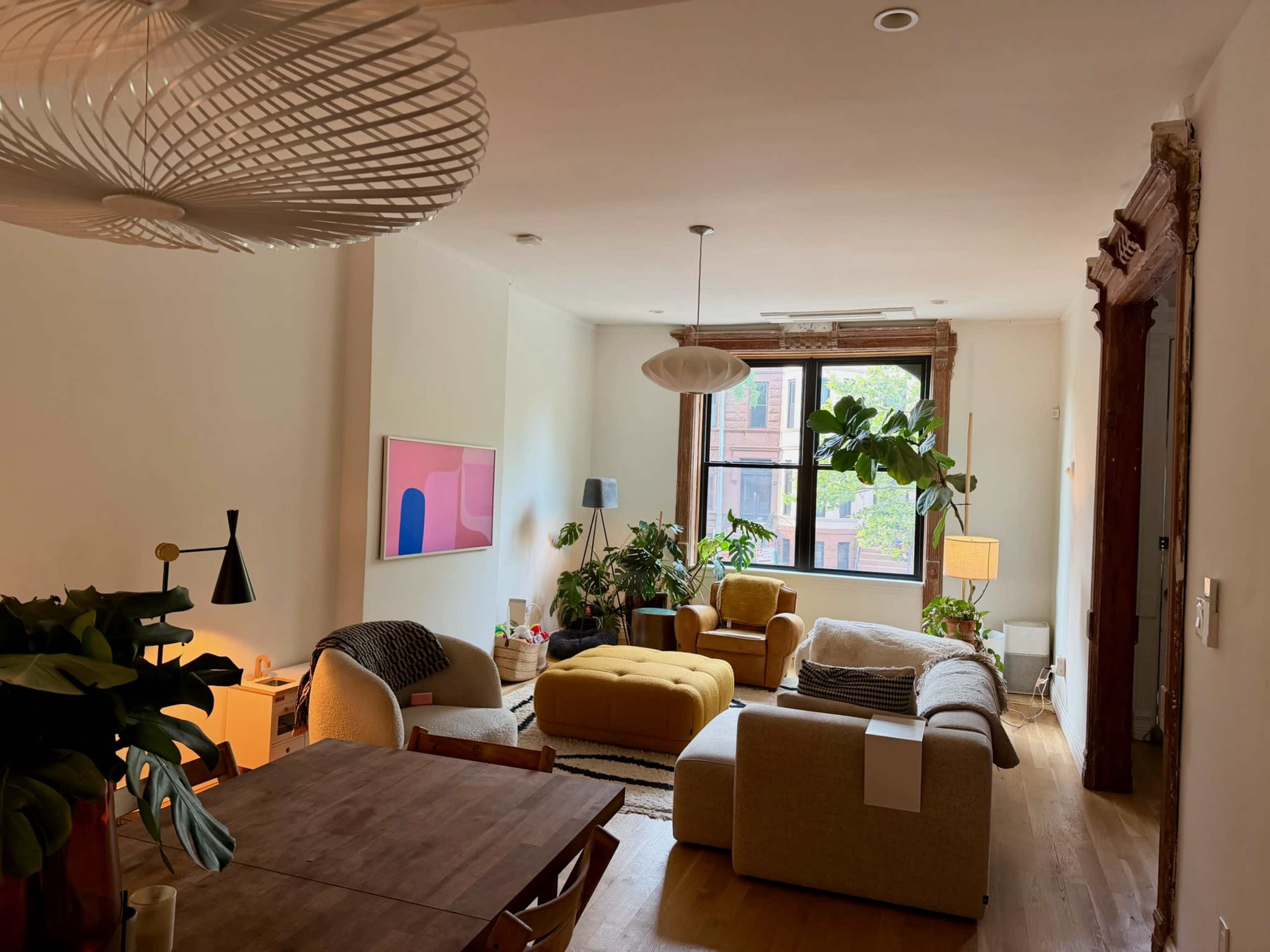 The image shows a bright, modern living room with a mix of furniture, including a yellow ottoman and several plants, illuminated by natural light from large windows.