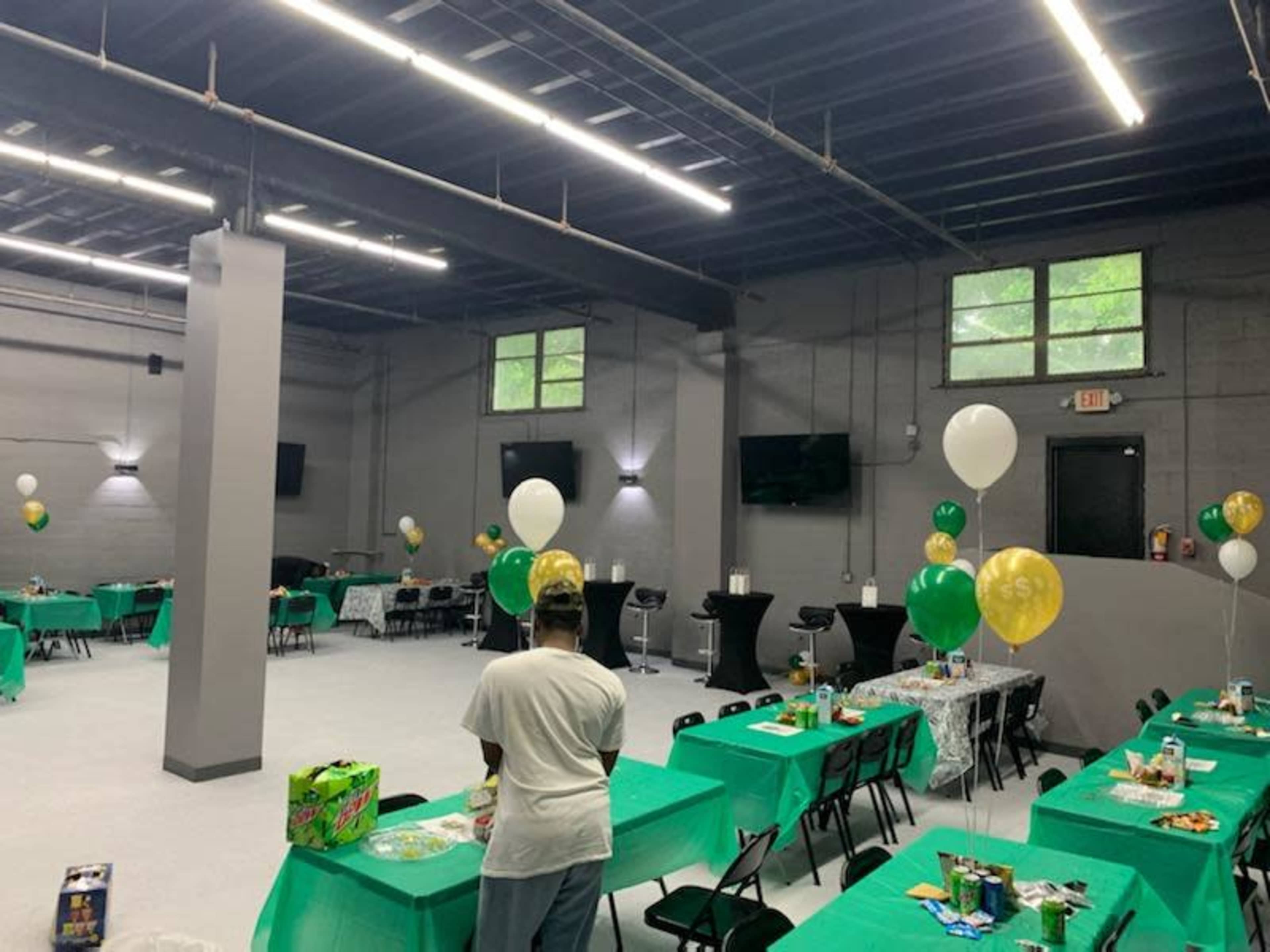 A spacious event room is set up with green and gold tablecloths, balloons, and various food items on tables for a gathering.