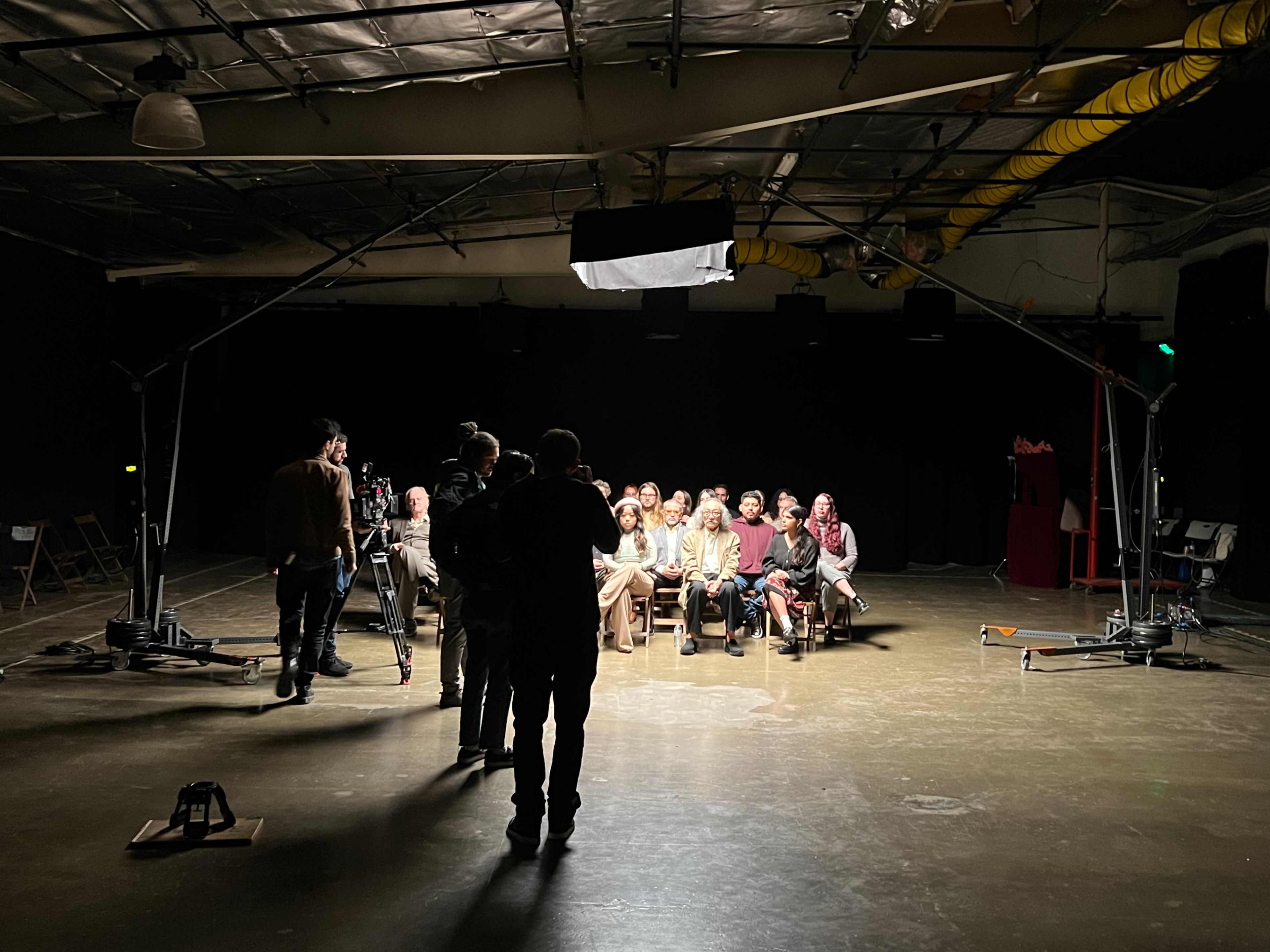 A group of seated individuals is facing a camera in a dimly lit studio, while crew members set up equipment around them.