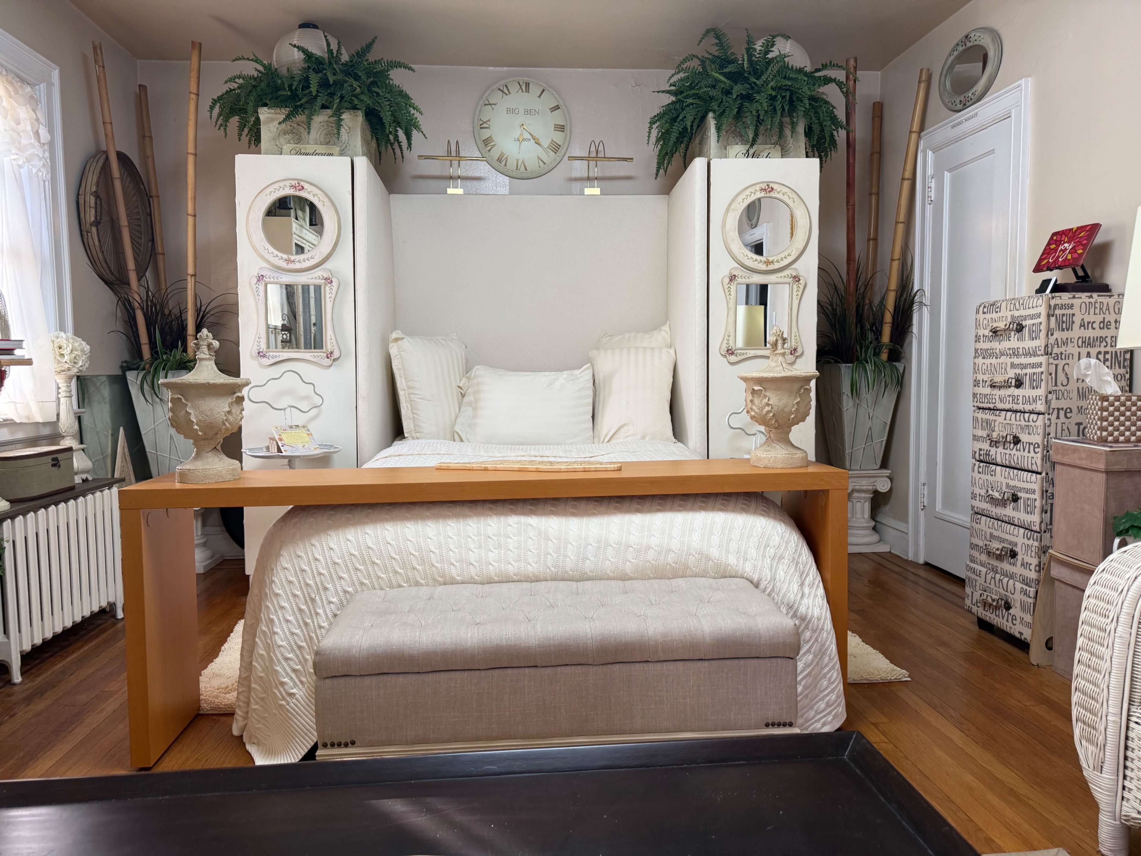 The image shows a cozy bedroom with a large bed flanked by decorative mirrors, plants, and a wooden table at the foot of the bed.