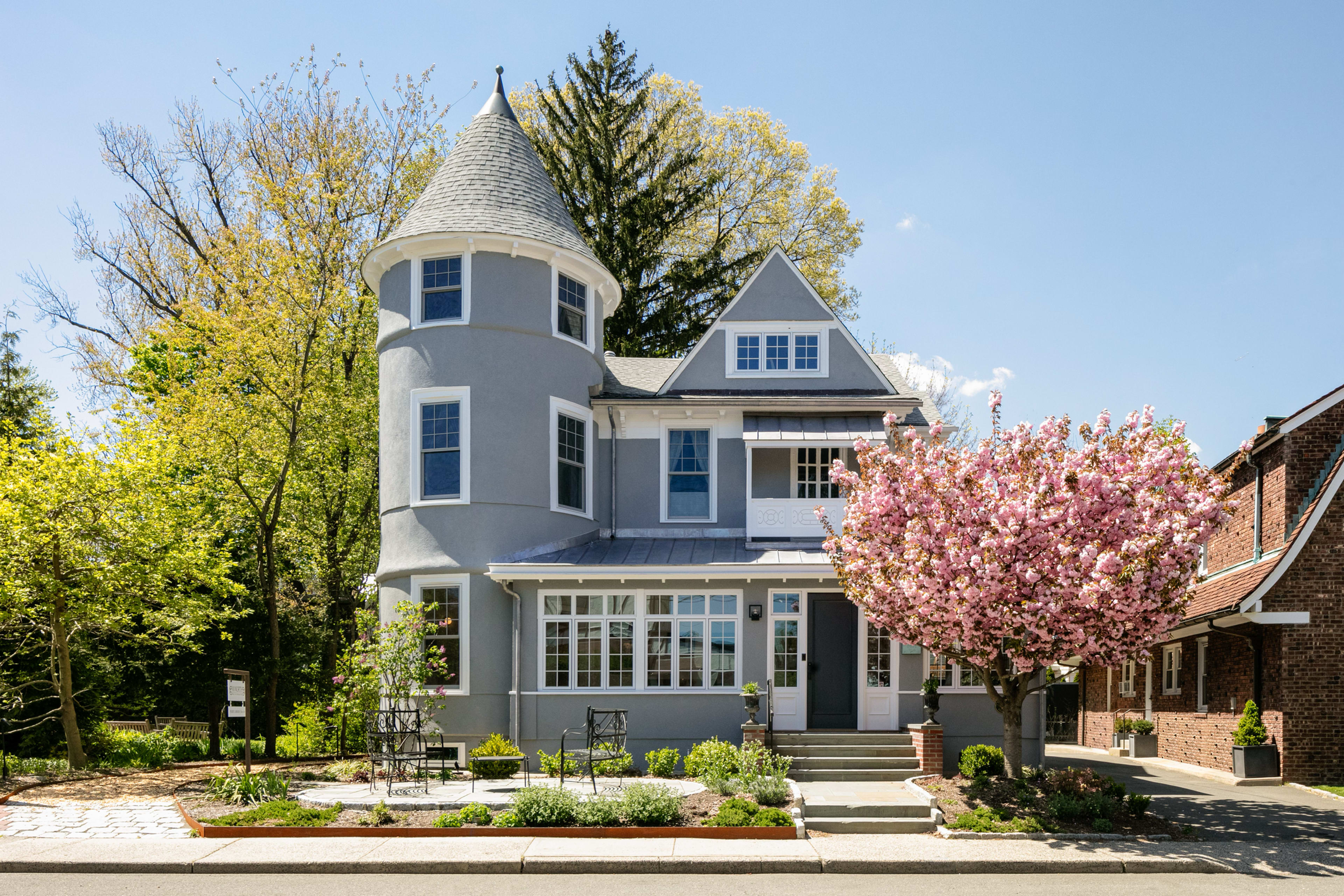 A gray house with a turret features a large front porch and a blooming cherry blossom tree in the yard.