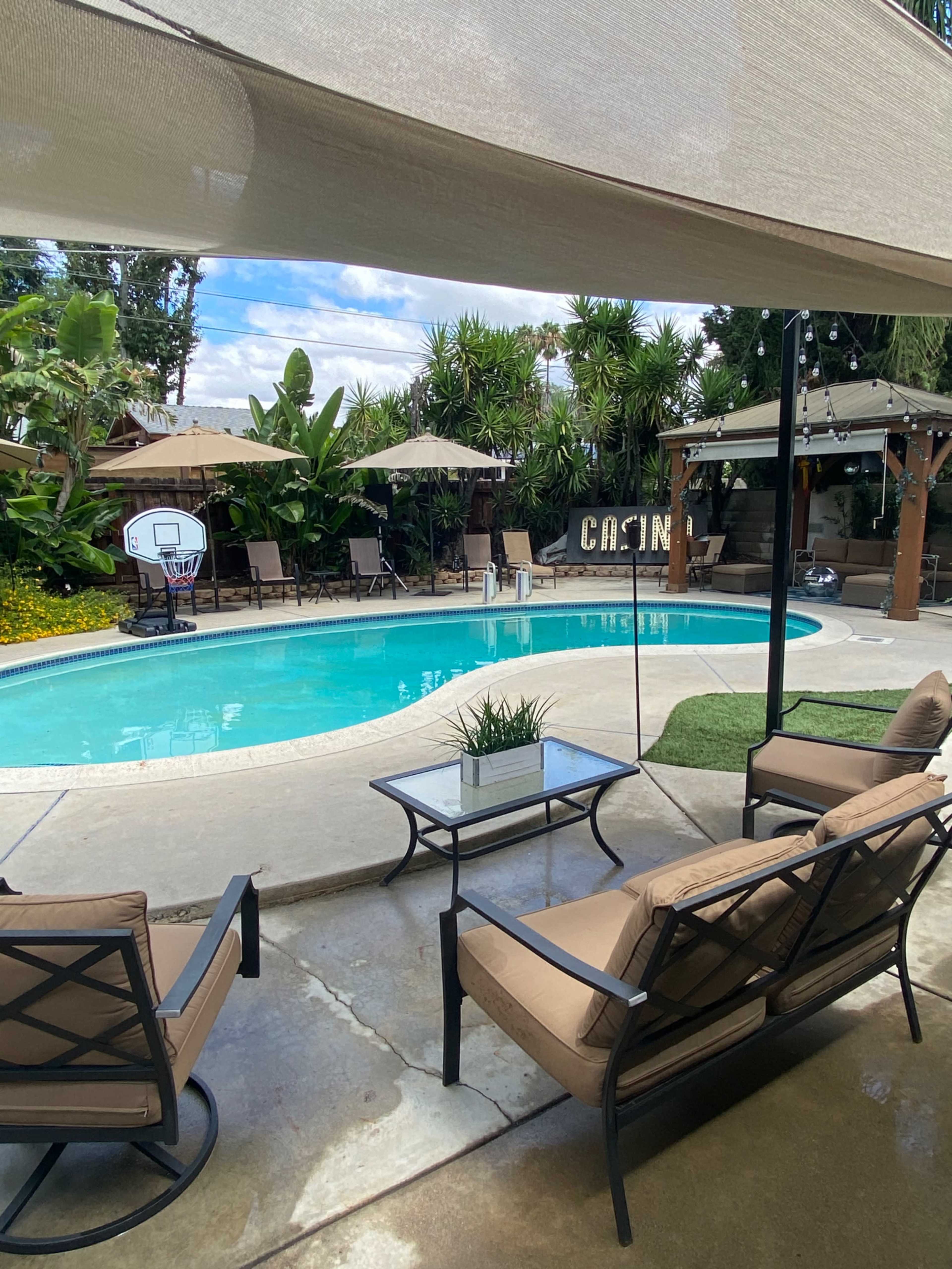 A seating area with two chairs and a glass table overlooks a swimming pool surrounded by palm trees and patio umbrellas.