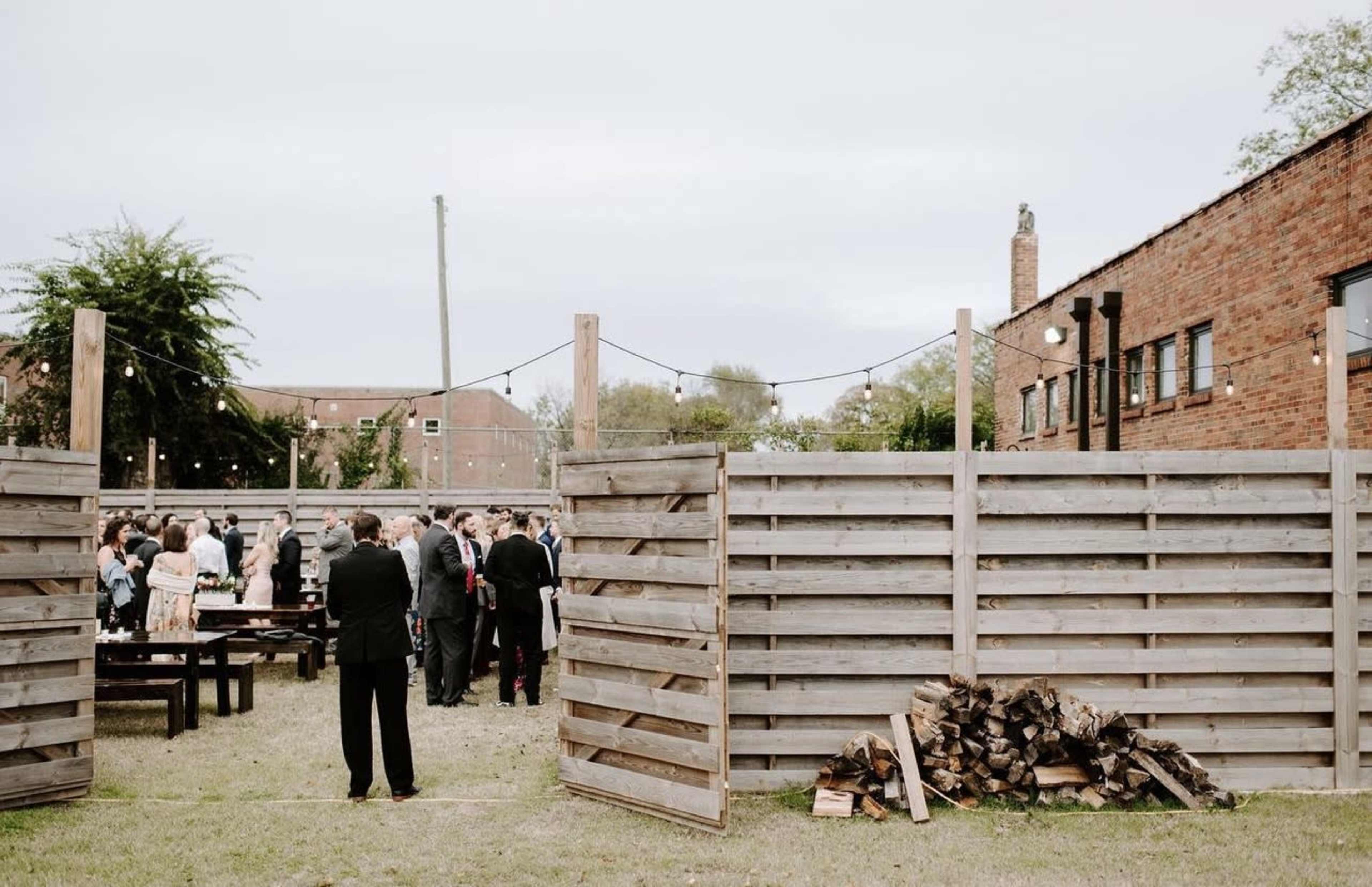 A gathering of people socializes in an outdoor area enclosed by wooden fences and illuminated by string lights.