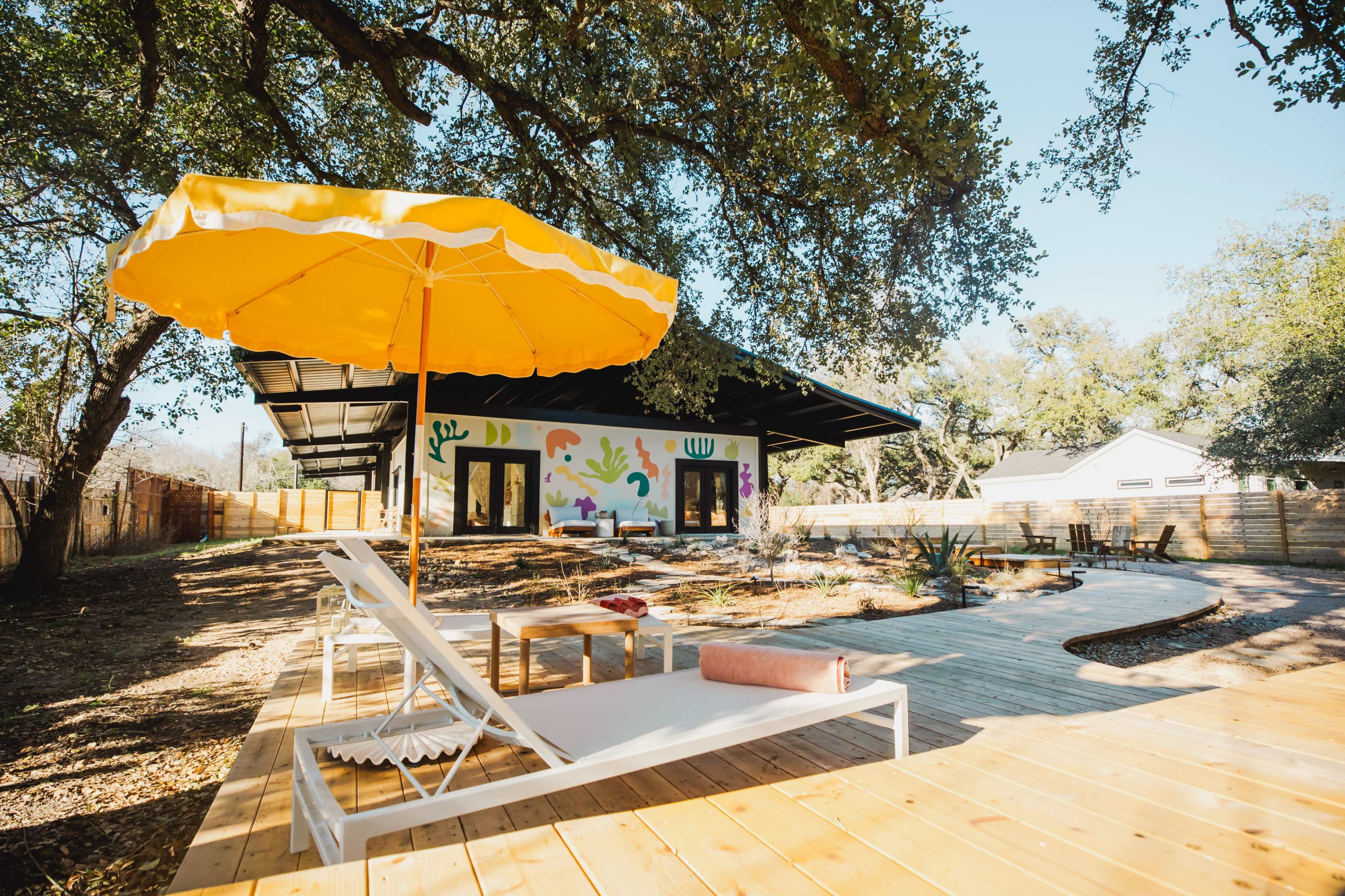 A vibrant outdoor space features a yellow umbrella shading a white lounger next to a wooden deck, with a modern house adorned with colorful murals in the background.