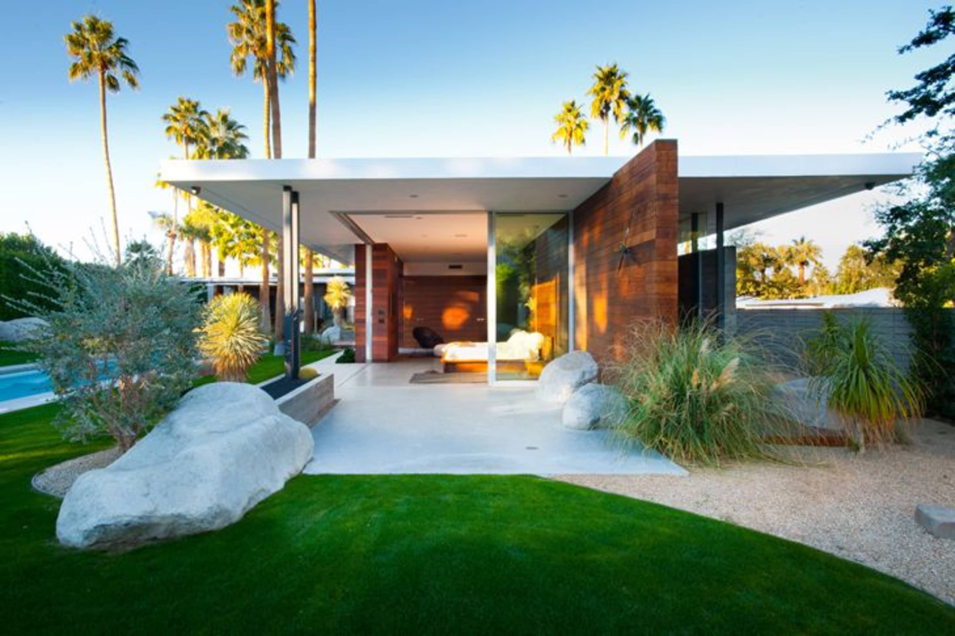 A modern single-story house with a flat roof is surrounded by lush greenery and large rocks, featuring a patio that opens to a swimming pool area.